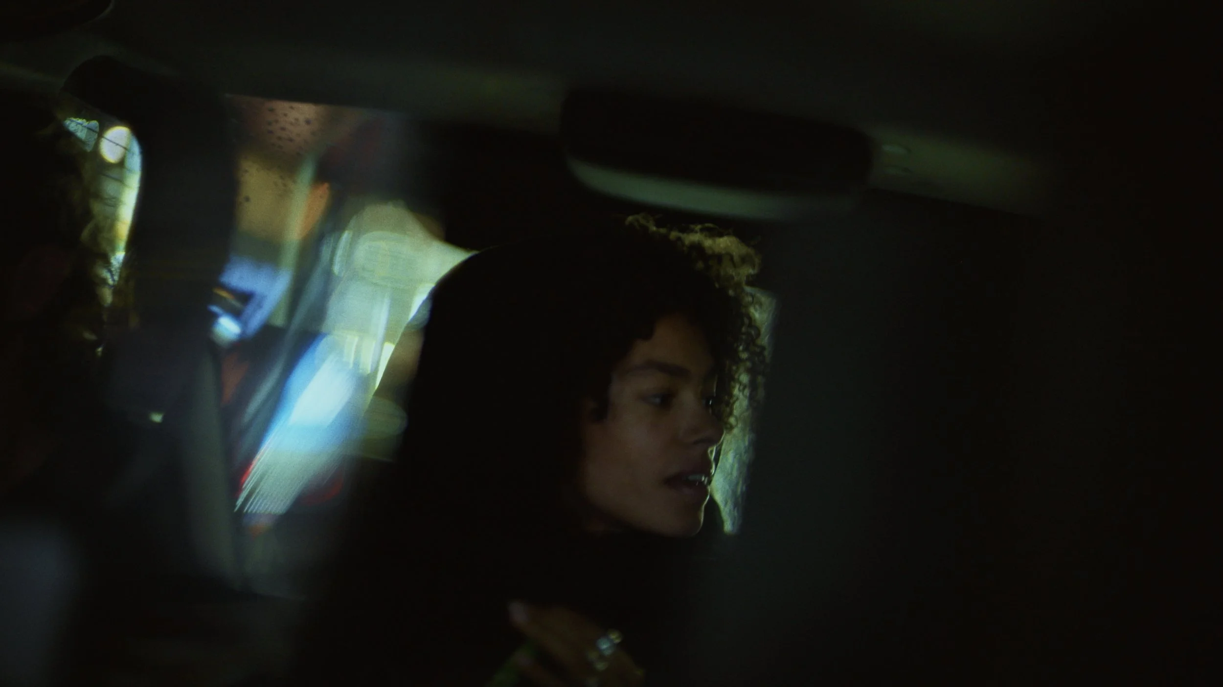 A woman with curly hair is in a dark environment, with reflections and light streaks around her, partially visible.