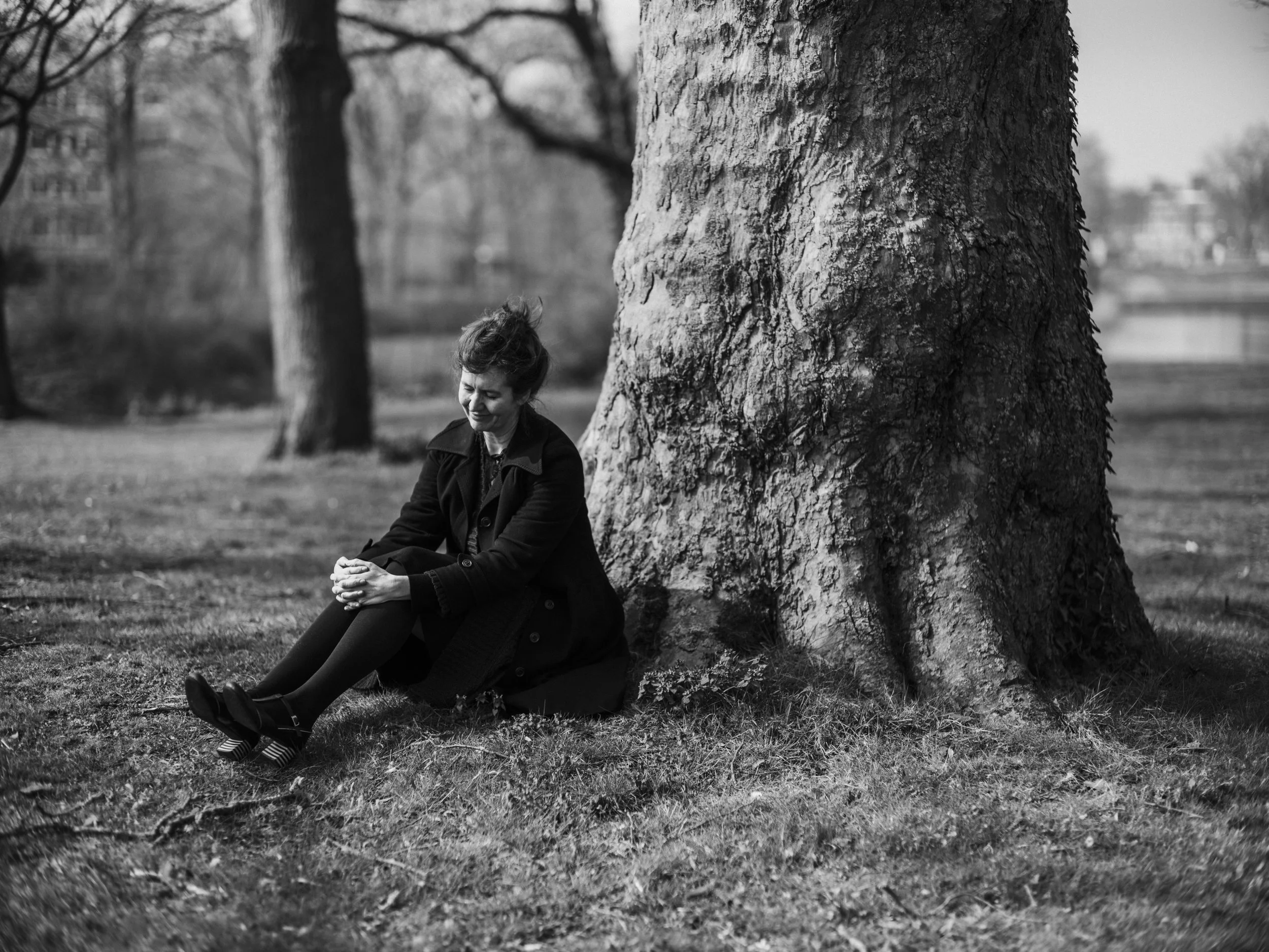 A woman with short hair sitting on the grass, leaning against a large tree, in a park.