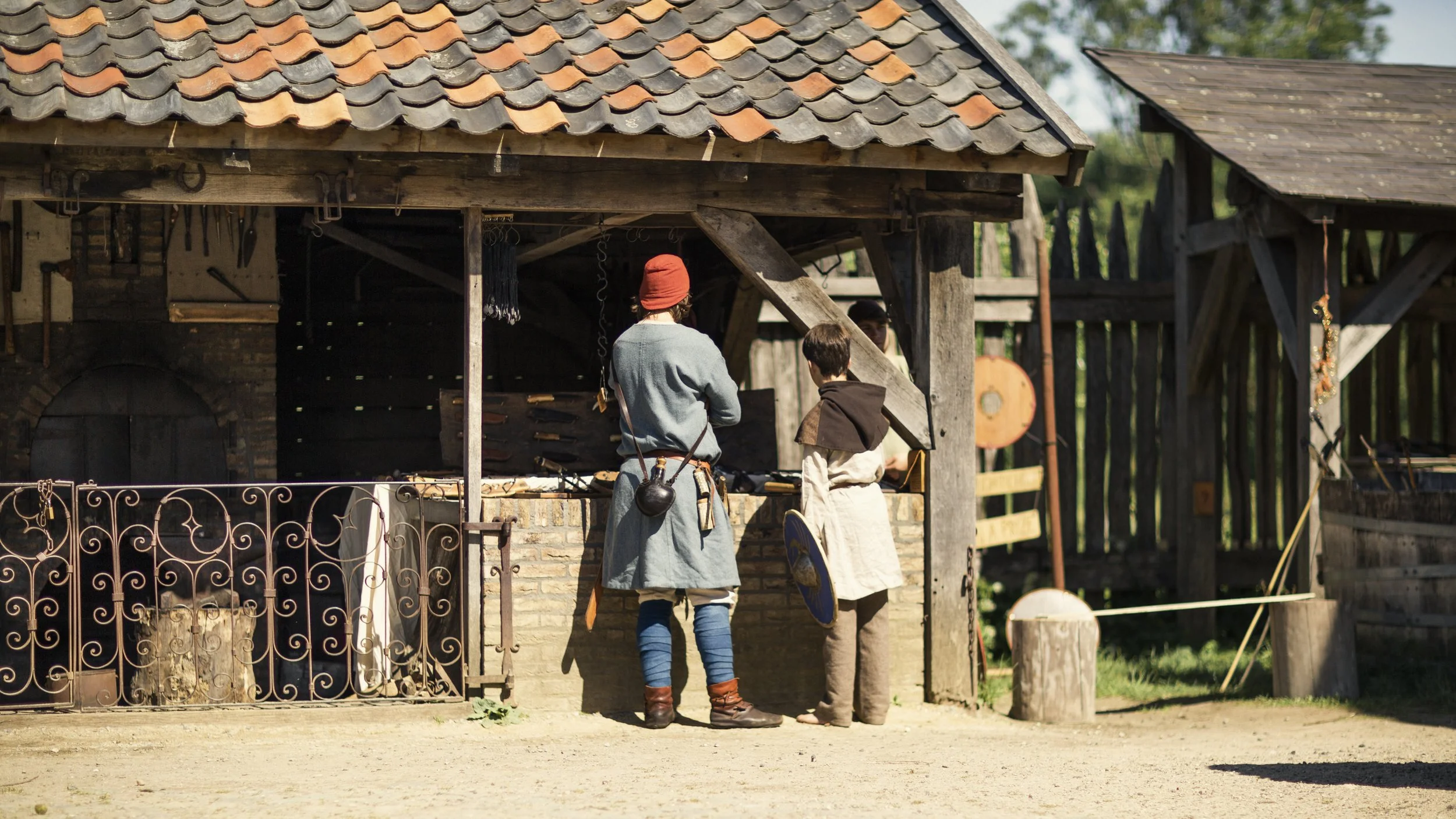 Two people dressed in medieval clothing stand outside a rustic blacksmith's workshop made of wood and stone, with a sloped tiled roof. They are engaged in conversation or observing something inside the workshop.