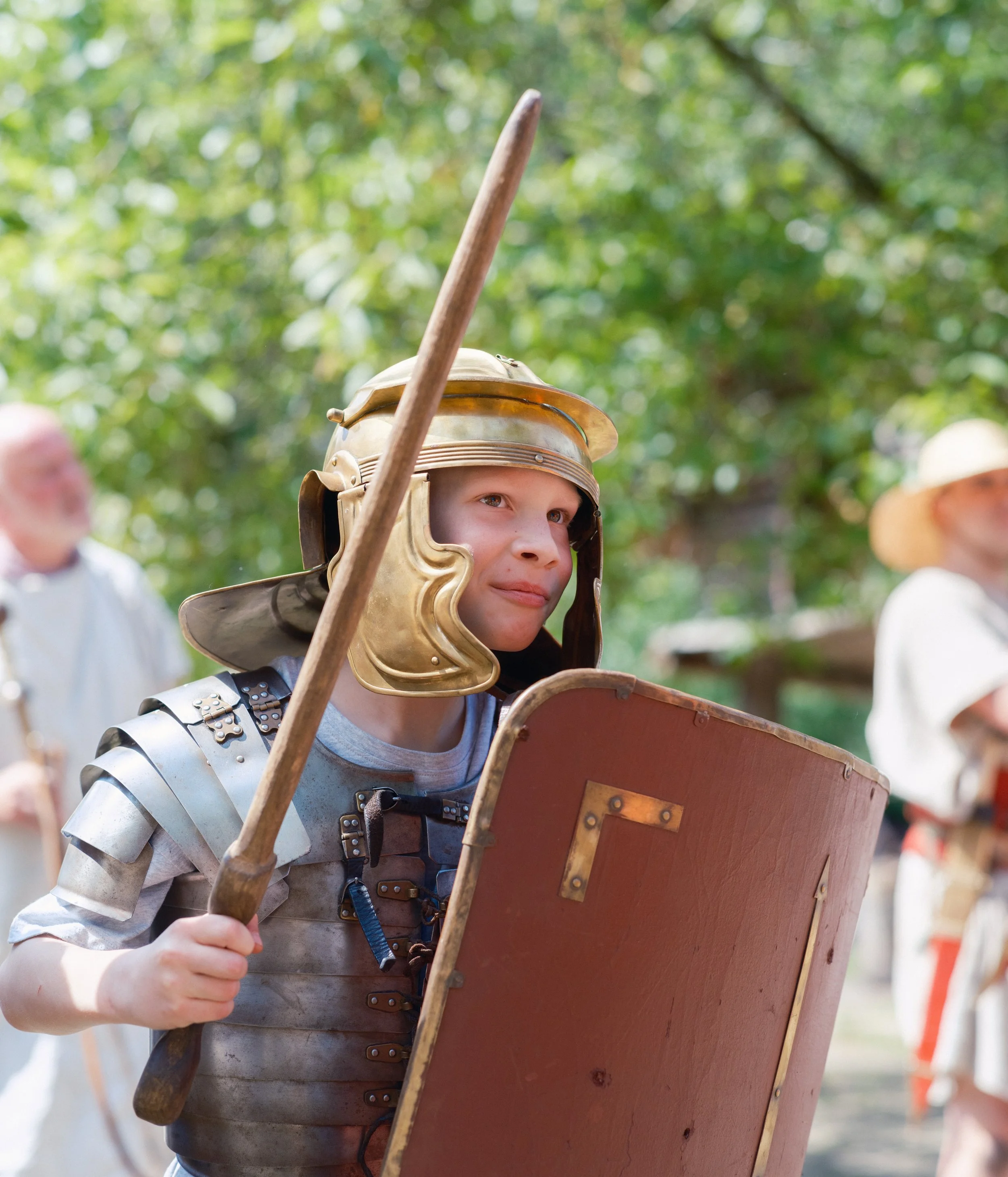 A young person dressed as a Roman soldier, wearing a metallic helmet and armor, holding a shield and a wooden spear, outdoors with green trees in the background.