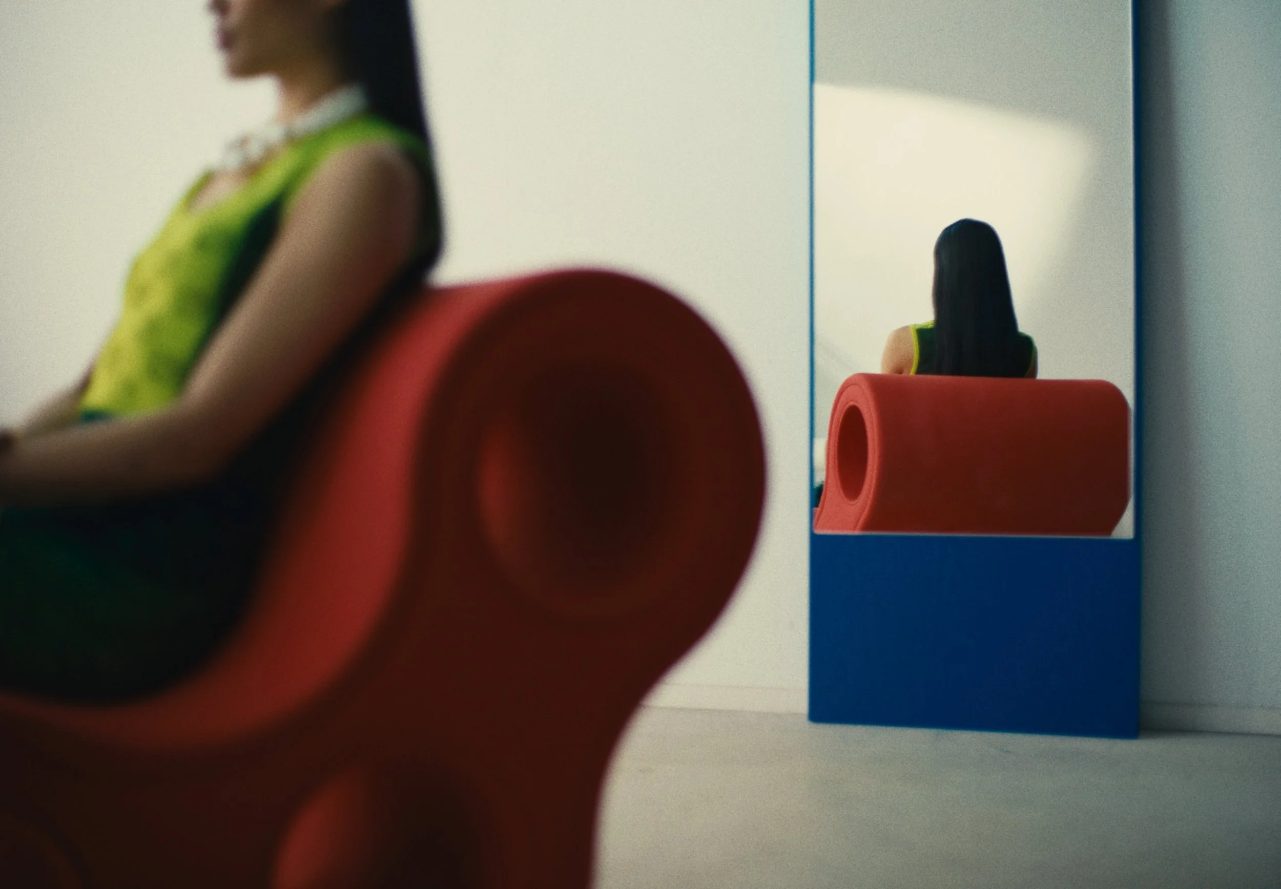 A blurred woman in bright clothing seated in front of a mirror with a red chair reflected behind her.