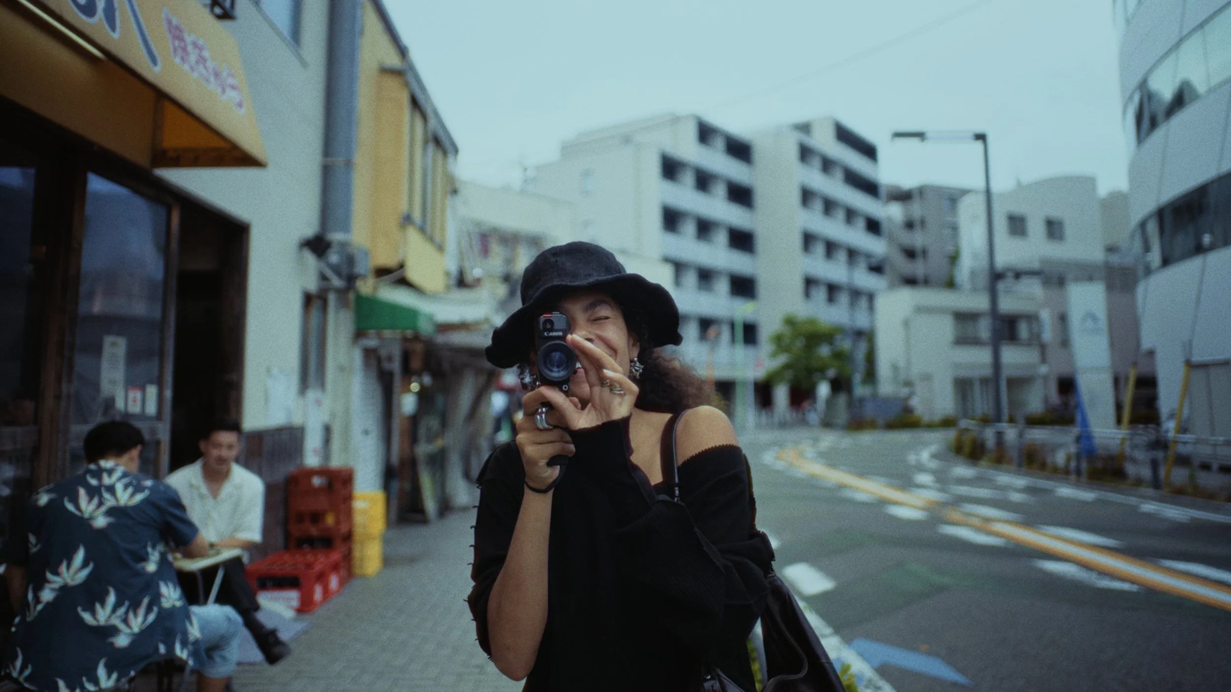 A woman with curly hair wearing a black hat and black clothing takes a picture with a camera on a city street with buildings and some people sitting at a table in the background.