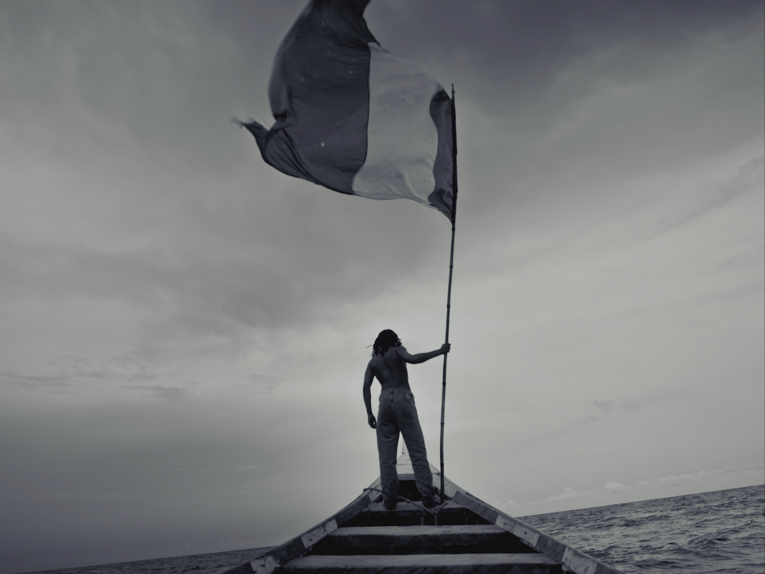 A person standing on a boat holding a flagpole with a black and white flag, against a cloudy sky and calm water.