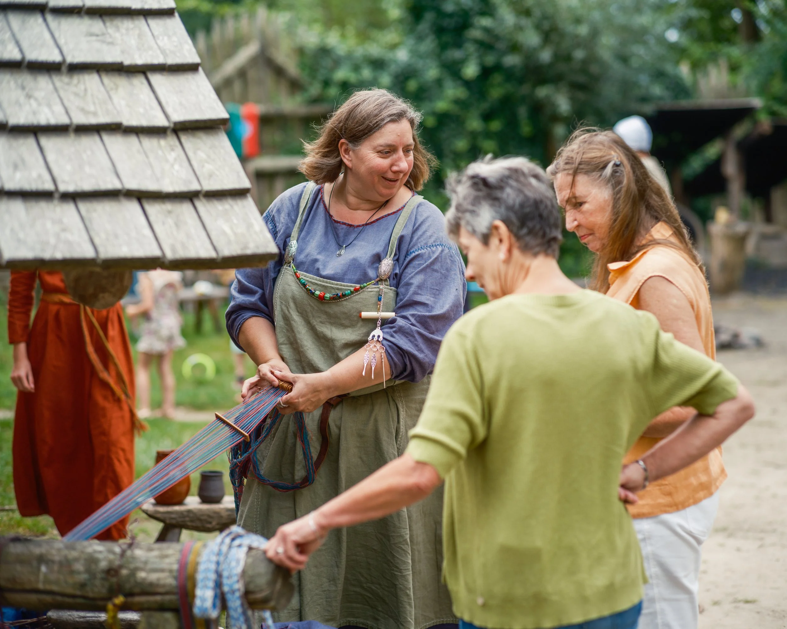 Group of people gathered outdoors, engaging in a craft or weaving activity, with trees and wooden structures in the background.