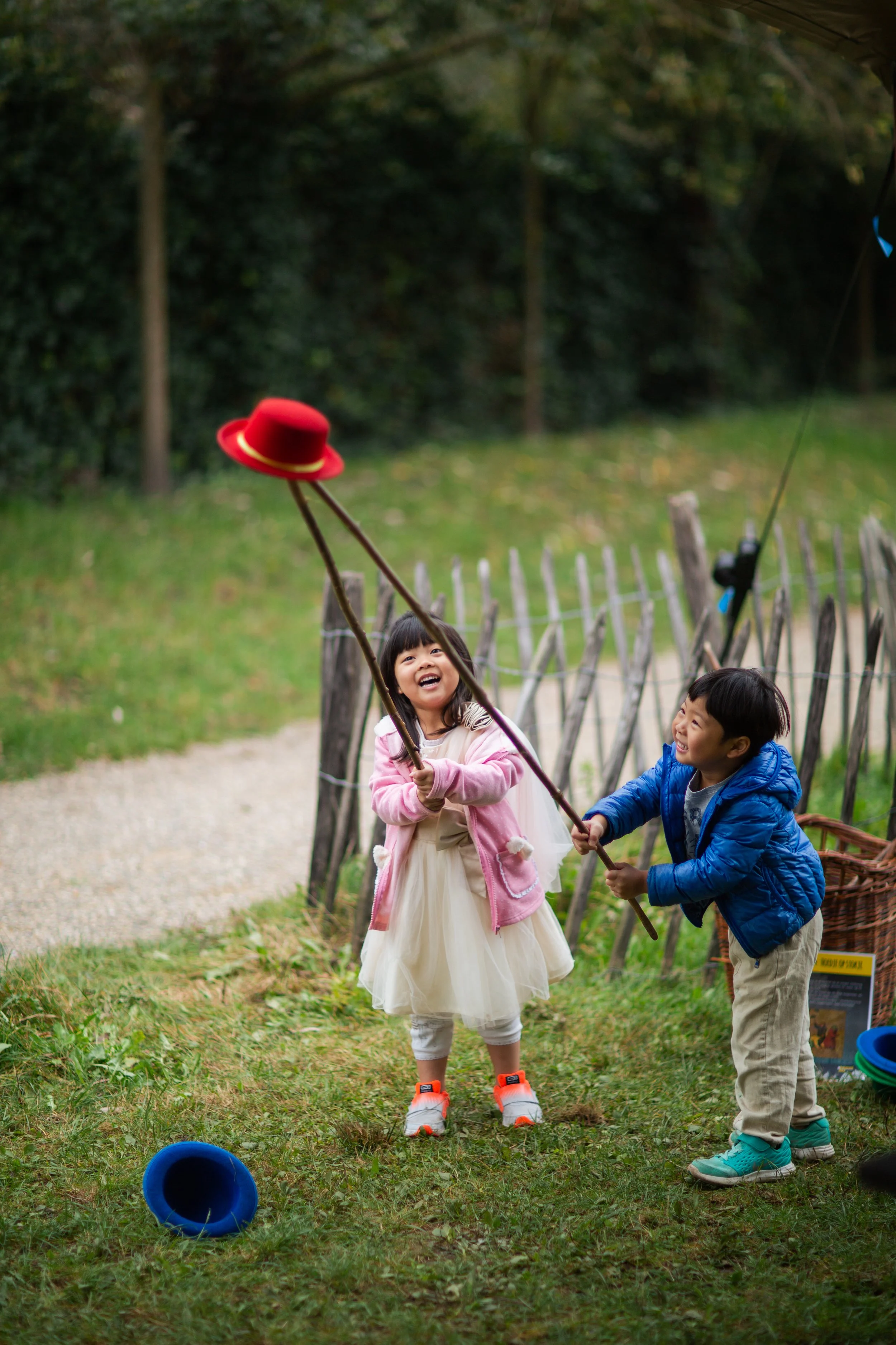 Two children playing a game of ring toss outdoors near a wooden fence, with trees in the background. The girl is wearing a pink hoodie and a white dress, and the boy is dressed in a blue jacket and beige pants, both laughing and holding a ring.