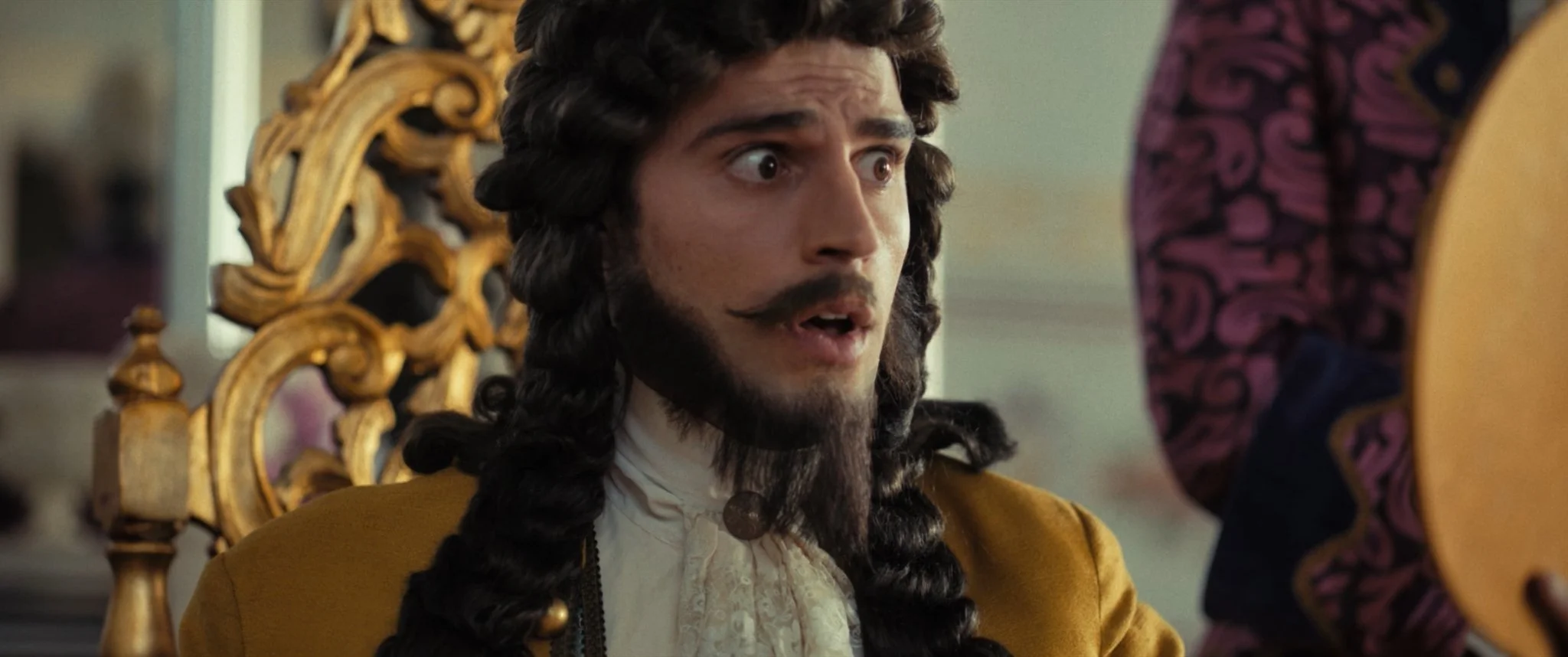 A man with long curly hair and a beard, dressed in historical costume, looking surprised or shocked, sitting on an ornate gold-colored throne.