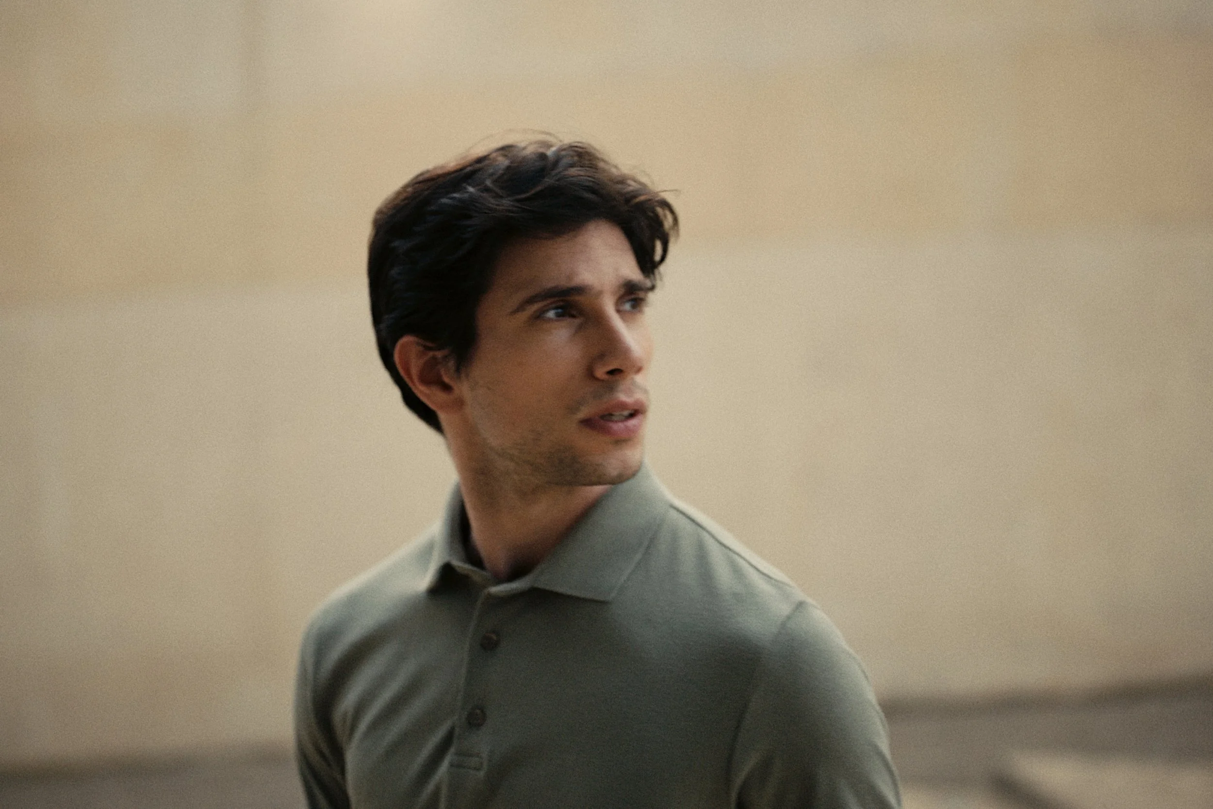 A young man with dark hair styled back, wearing a light-colored button-up shirt, looking to his side against a plain, beige background.