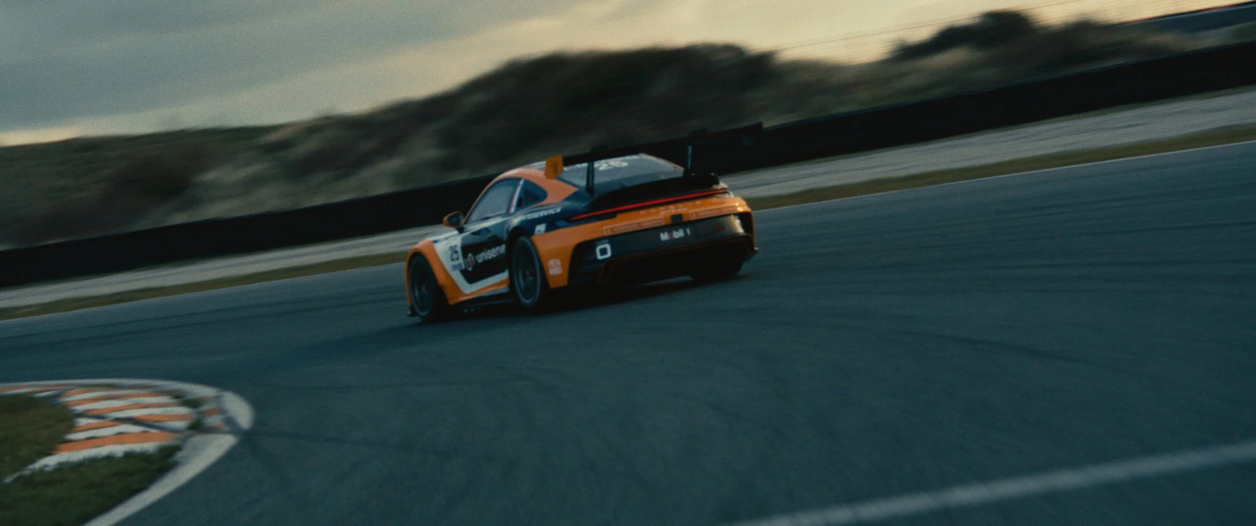 An orange and black race car speeding on a race track during dusk or dawn.