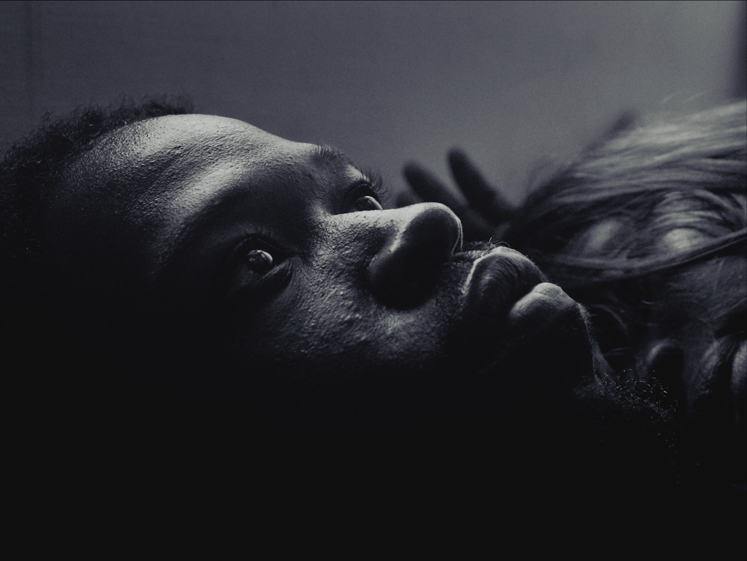 A woman with dark skin lying on her back, gazing upward with wide eyes, with her hand gently touching her face and long hair spread out beside her, in a black and white photograph.