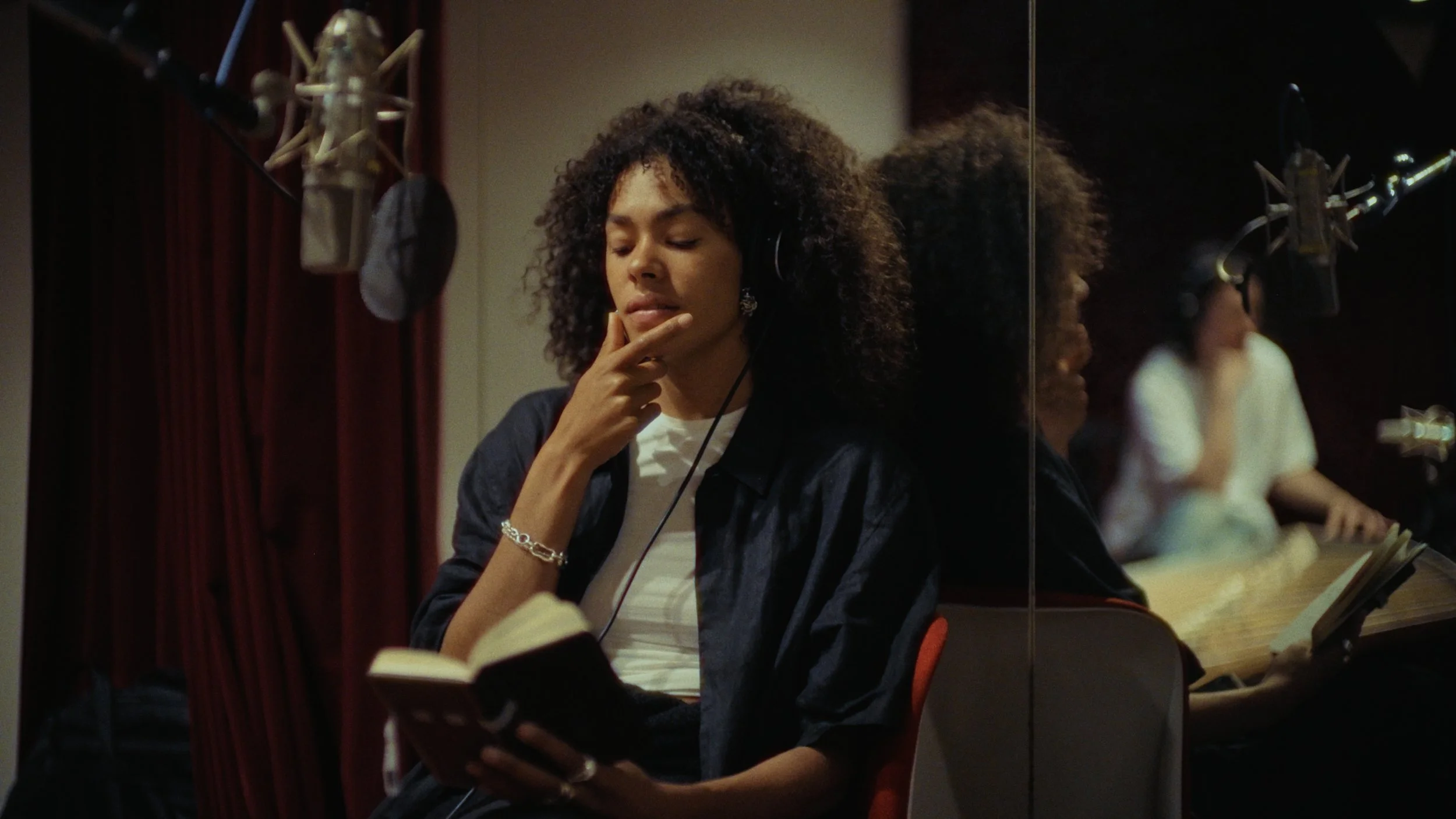 A woman with curly hair wearing headphones, reading a book, and holding her chin, standing in a recording studio near a mirror.