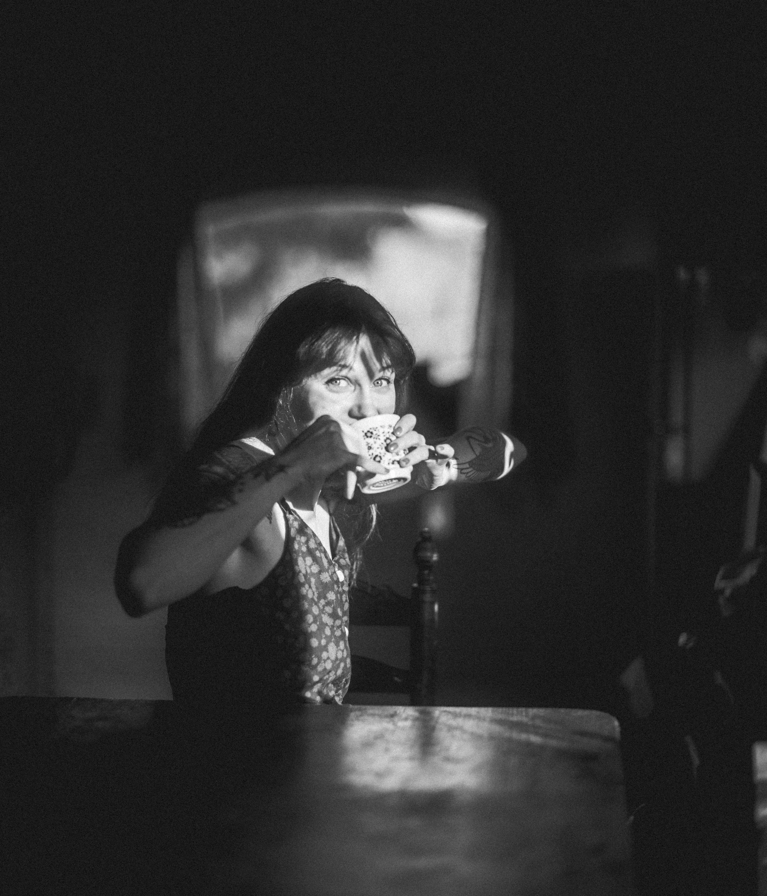 A woman with long dark hair and light skin is sitting at a table, holding a cup with both hands, and looking directly at the camera in a dimly lit room.