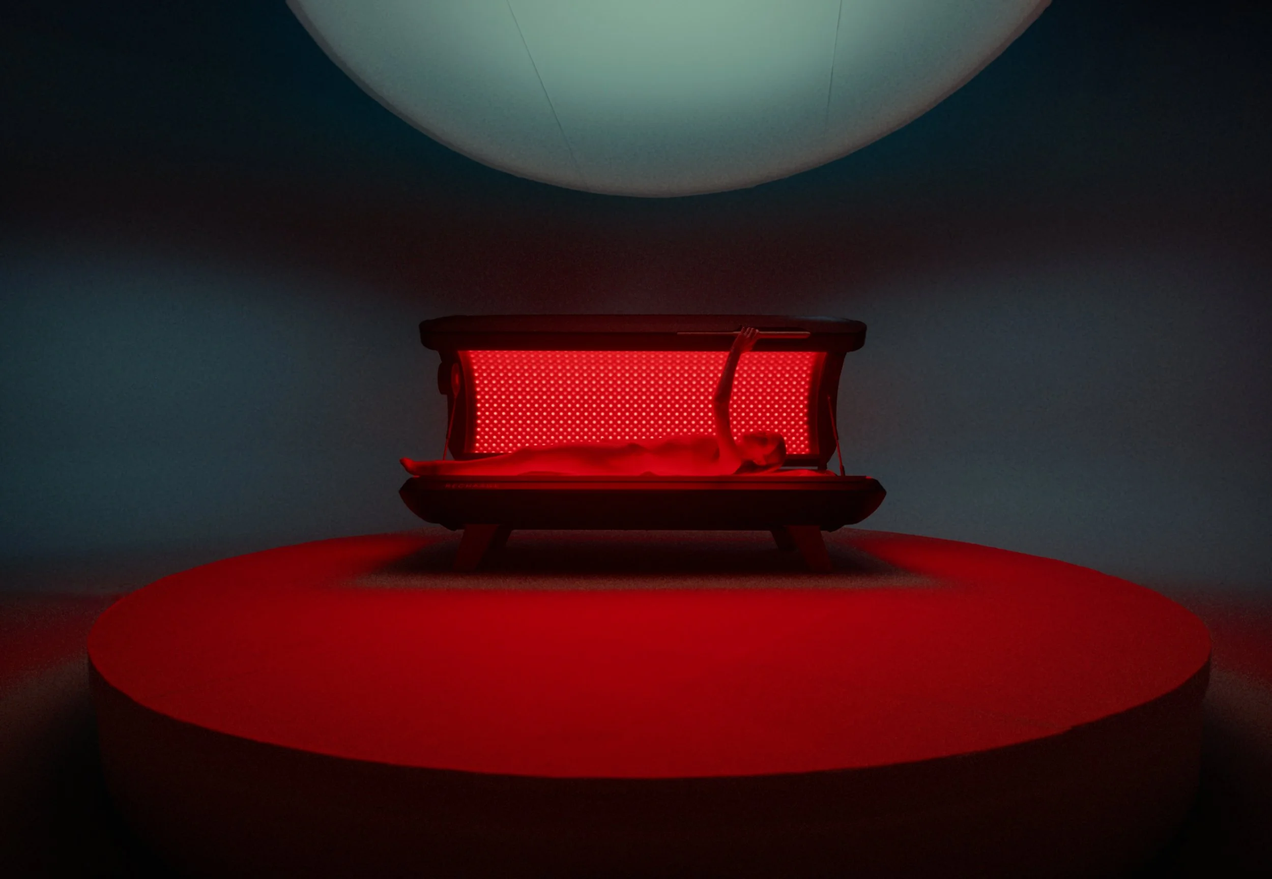 A red bench with a person lying on it, positioned beneath a large spherical object in a dark space with a red circular surface underneath.