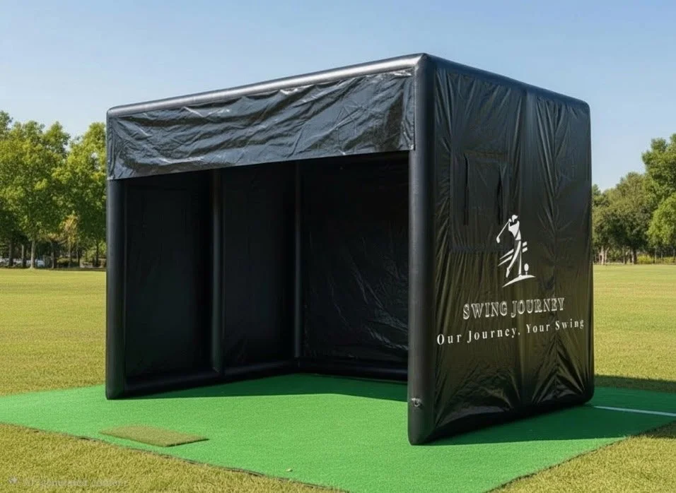 Black golf simulator tent with brand name Swing Journey, set up on a green turf in an outdoor field with trees in the background.
