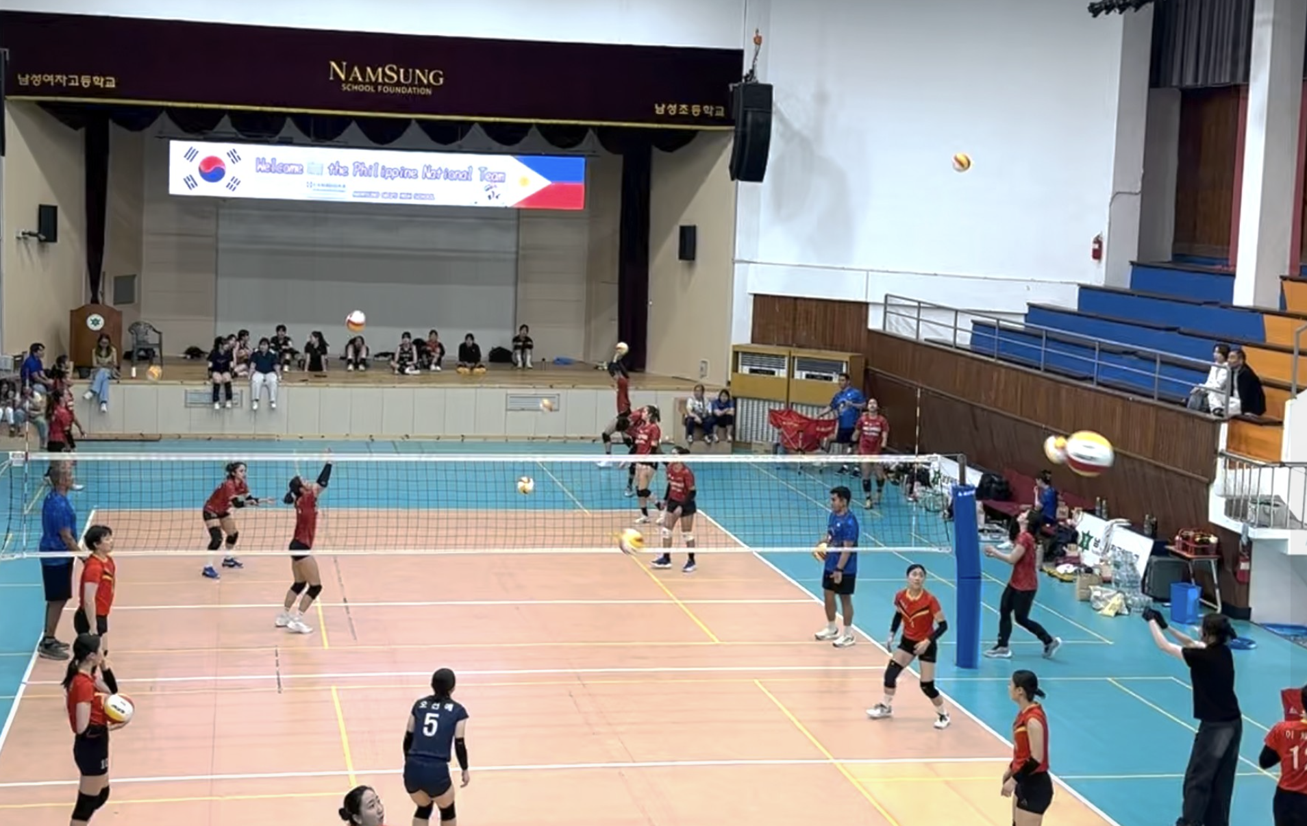 Indoor volleyball match with players in red and blue uniforms on the court, some jumping to hit the ball, and others preparing to receive. Spectators and team members are seated or standing around the court, with a scoreboard and banners visible in t