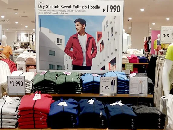 Display of various colored dry stretch sweat full-zip hoodies in a store, price marked at 1,990 pesos, with a large poster of a man wearing a red hoodie.