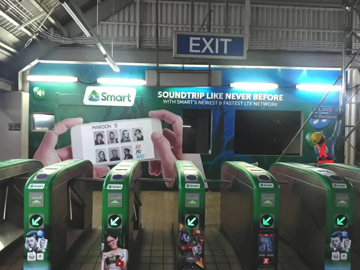 Underground train station with turnstiles, an advertisement for Smart cellular service on the wall, and a figure with a yellow helmet and red safety vest standing on the right side.