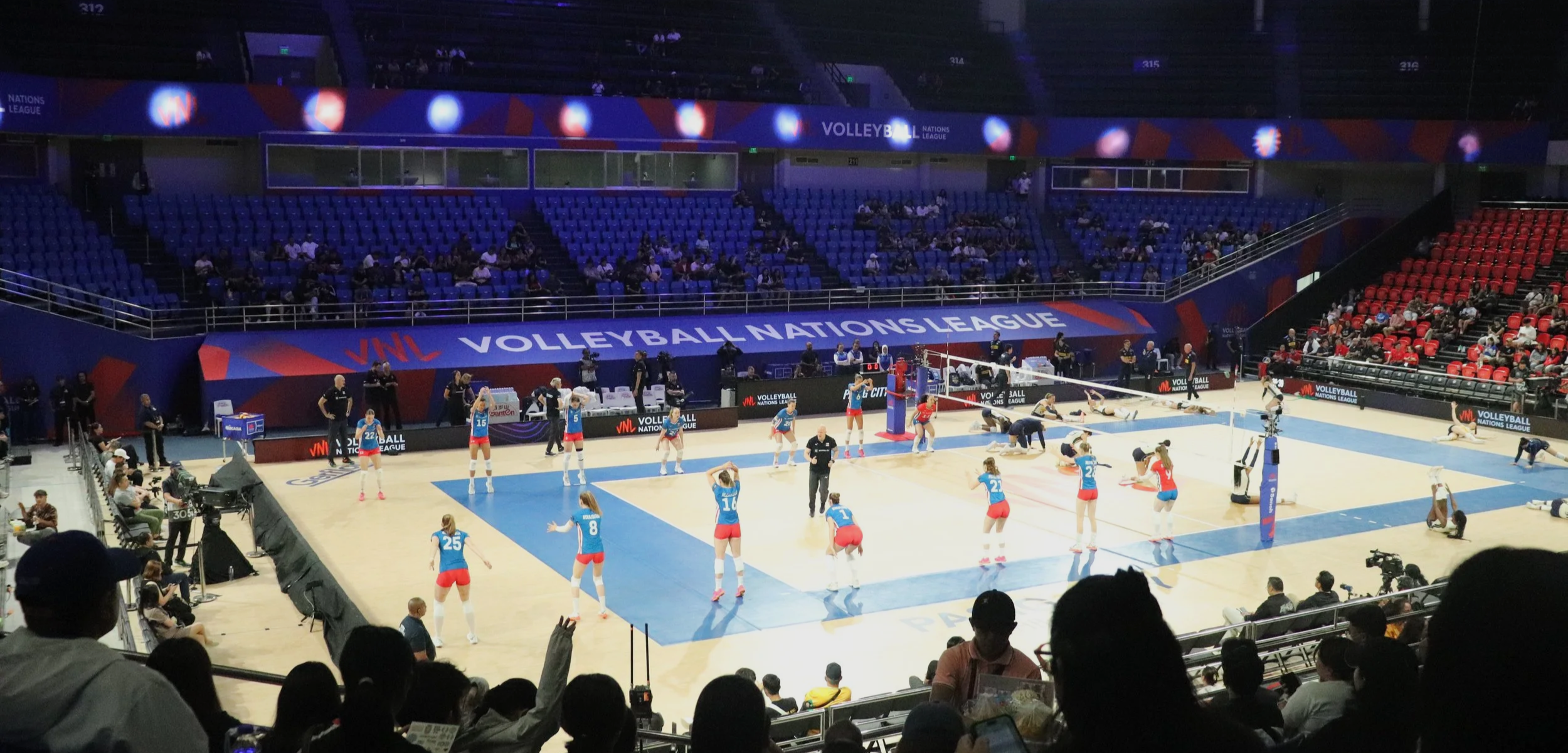 An indoor volleyball game on a court with players in blue and red uniforms, some players diving and falling, with audience seating and a scoreboard visible.