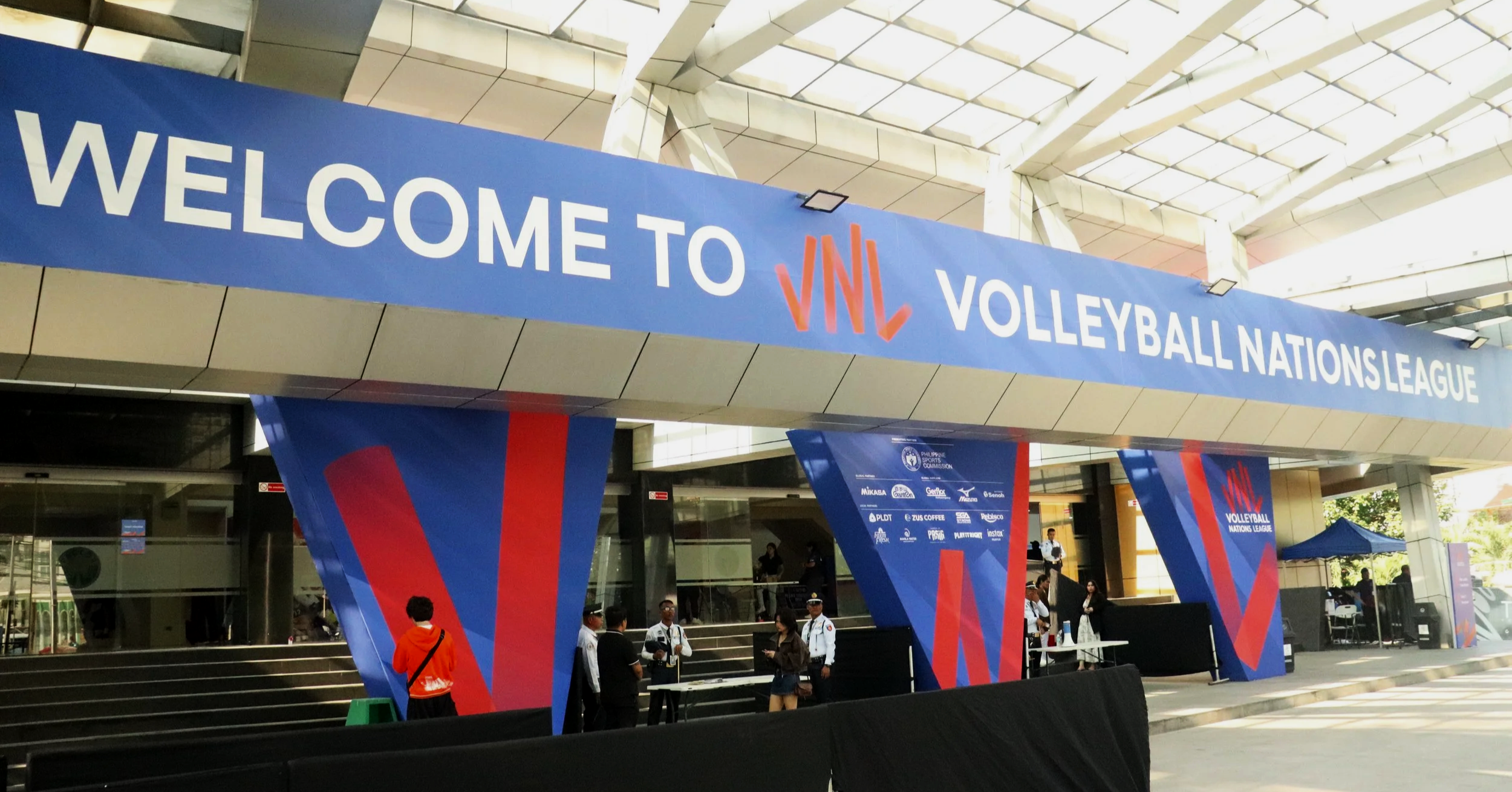 Entrance to volleyball event at the Nations League with a large blue banner reading "Welcome to VNL Volleyball Nations League" and people gathered underneath.