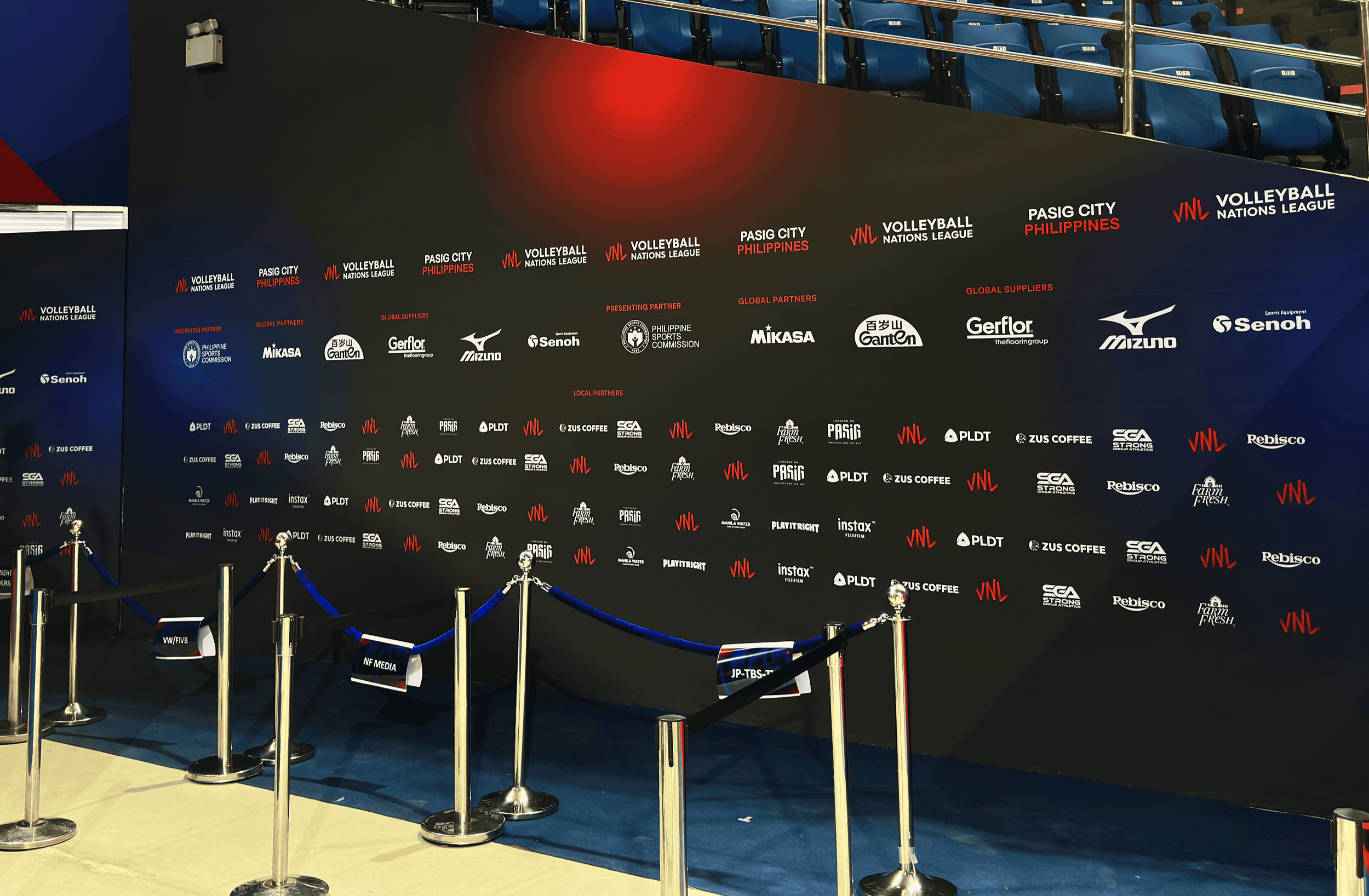 A volleyball arena with a black wall featuring logos of sponsors such as Pasig City, Philippines, Volleyball Nations League, Mikasa, Mizuno, and other brands. Blue seats are visible in the background, and a rope barrier sectioned off the area with sm