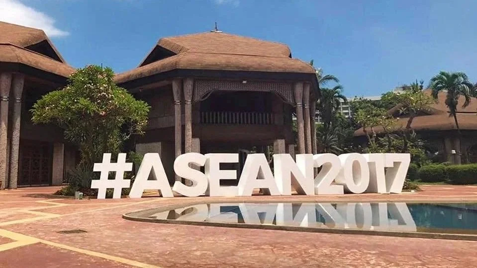 Large white hashtag sign reading #ASEAN2017 in front of traditional style building with trees and pool reflecting the sign, under a blue sky.