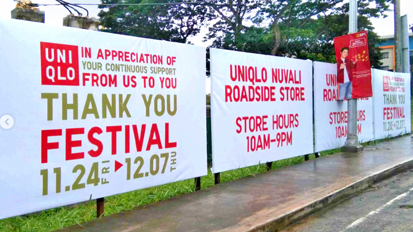 Set of large UNIQLO outdoor banners with red and green text on a white background, thanking customers during a festival from November 24 to December 7, alongside store hours and location details for UNIQLO Nuvali roadside store, with a sidewalk, a ut