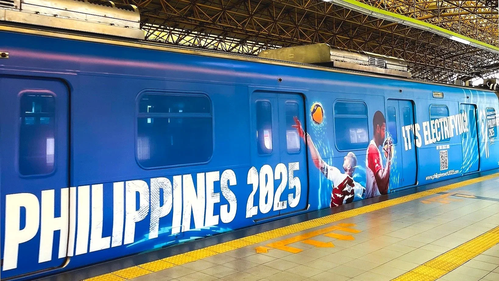 A blue train at a station with a banner that reads 'Philippines 2025' and shows athletes engaging in sports, with the slogan 'It's Electrifying' and a QR code.