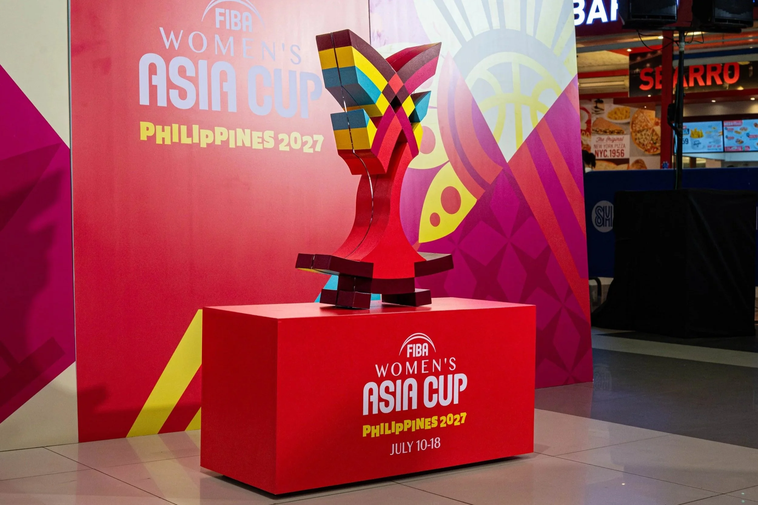 A large red 3D build up of the  FIBA Women's Asia Cup Philippines 2027 logo, scheduled for July 10-18. The stand has a colorful trophy sculpture and is set in an indoor venue with sound speakers and restaurant signs visible in the background.