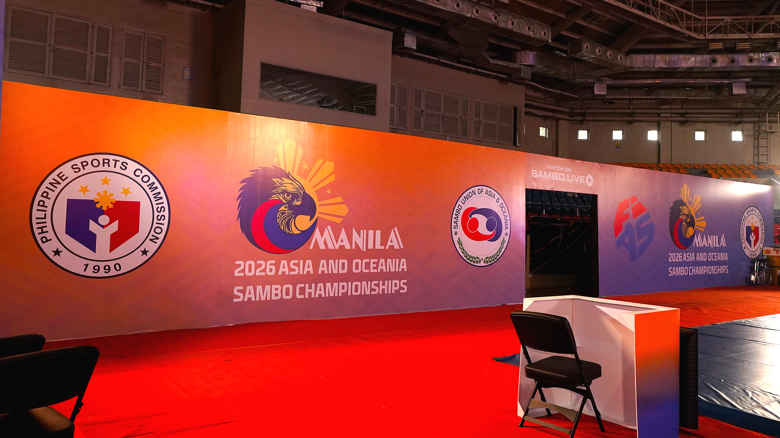 Indoor sports arena with a large orange banner displaying logos for the Philippine Sports Commission and the Sambo Union of Asia & Oceania, promoting the 2026 Asia and Oceania Sambo Championships in Manila. The arena has seating for spectators, an el