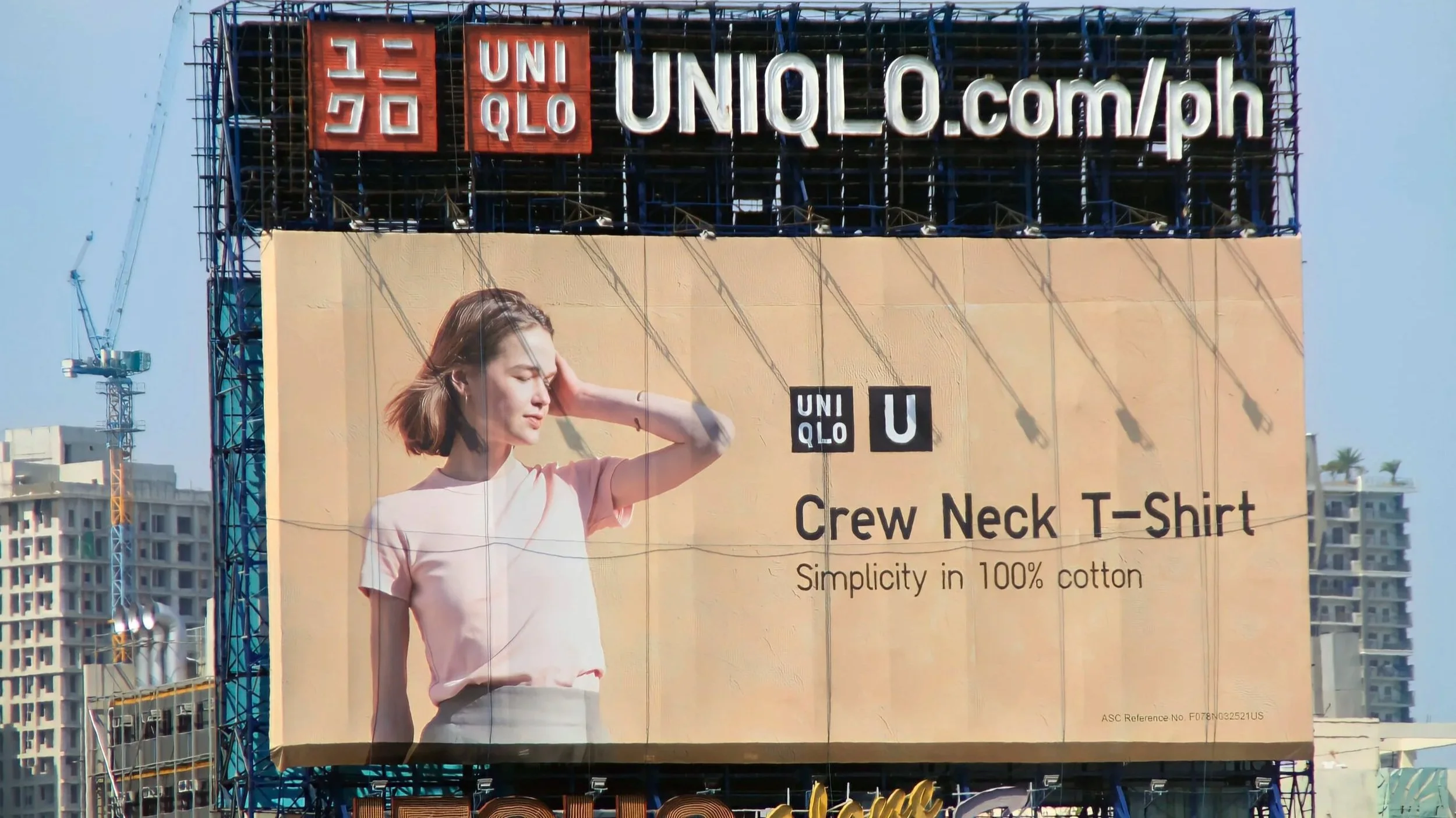 A billboard advertisement for Uniqlo featuring a woman in a pink Crew Neck T-Shirt. The billboard includes the Uniqlo logo, the website uniqlo.com/ph, and text highlighting the T-shirt's simplicity and 100% cotton material. The background shows a cit