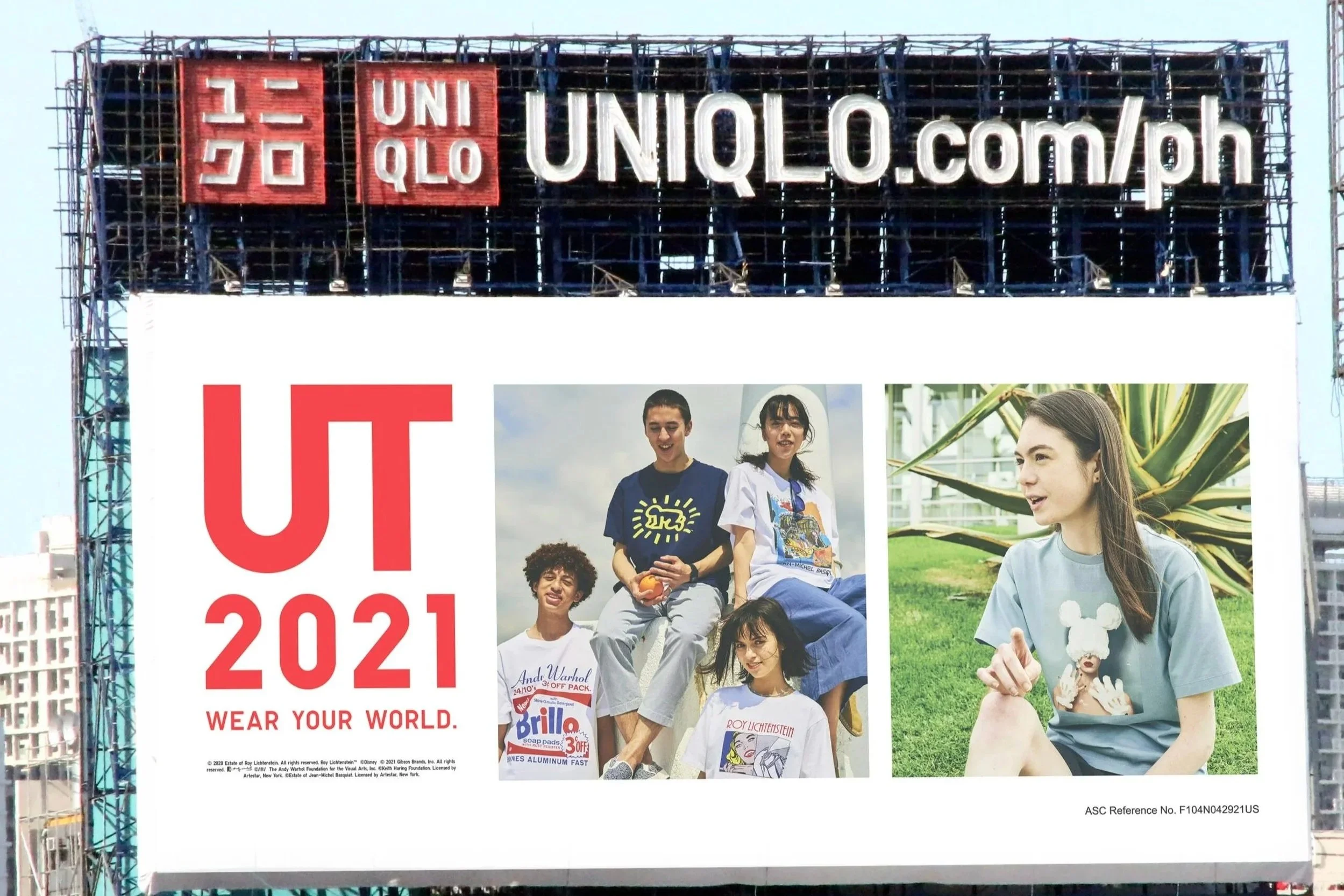 A billboard with a large sign at the top displaying the UNIQLO logo and website. Below the sign, there are photos of several young people outdoors, some sitting and some standing, wearing casual clothing, with a clear sky and city buildings in the ba
