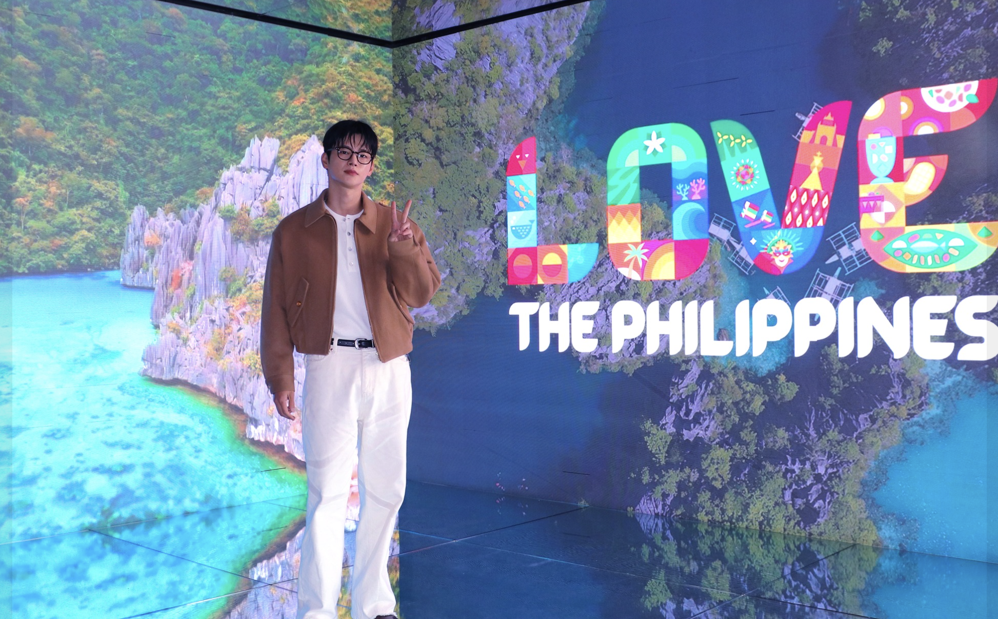 Korean singer and actor SEO INGUK, Philippine Tourism Ambassador to South Korea, wearing glasses, a brown jacket, a white shirt, and white pants standing in front of a large colorful sign that reads "LOVE THE PHILIPPINES" with tropical landscape imag