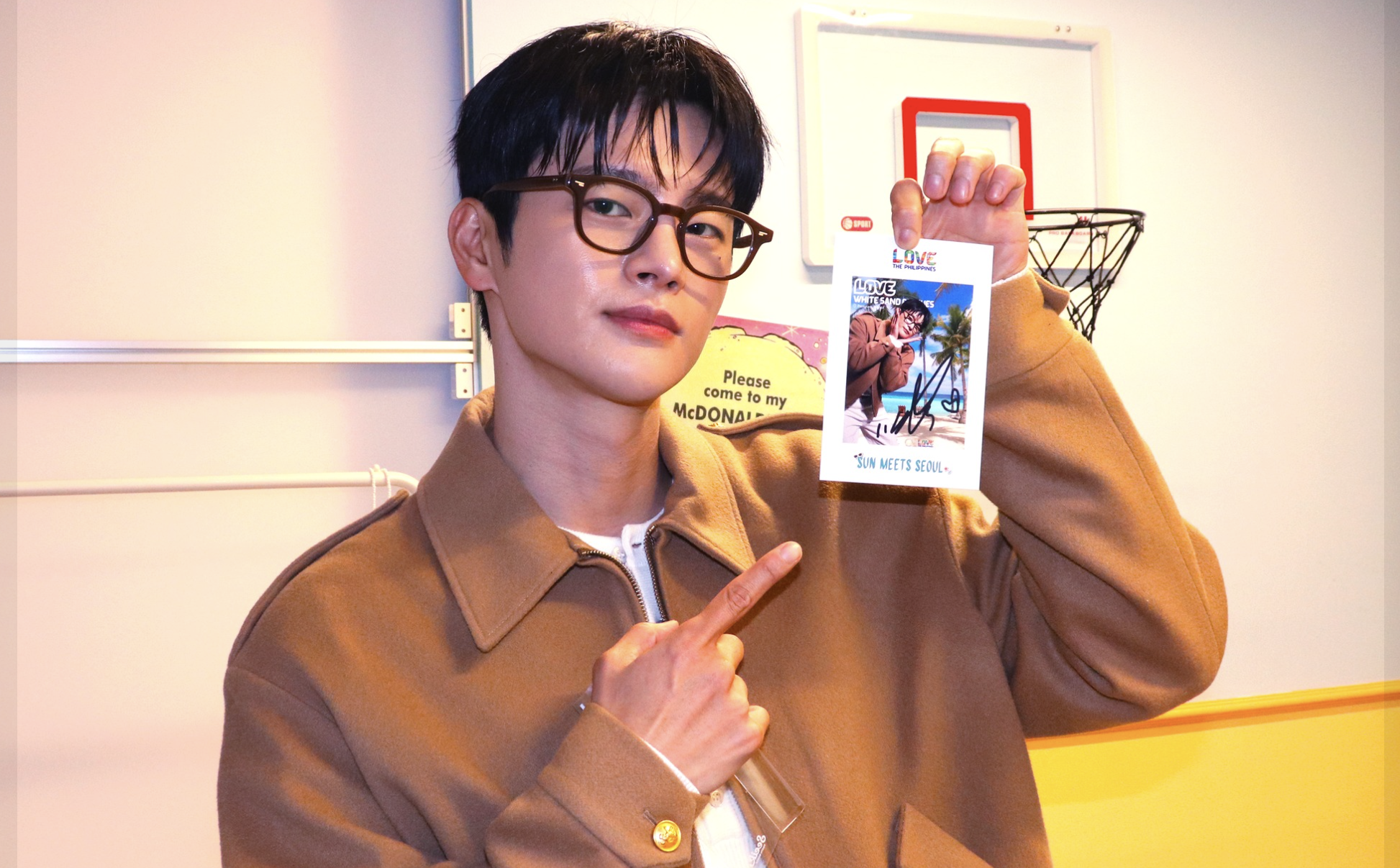 A young man with dark hair and glasses smiling while holding a signed photo at an indoor event. He wears a brown jacket and is pointing to the sign with his left hand. The background features a red-bordered basketball hoop and colorful wall decoratio