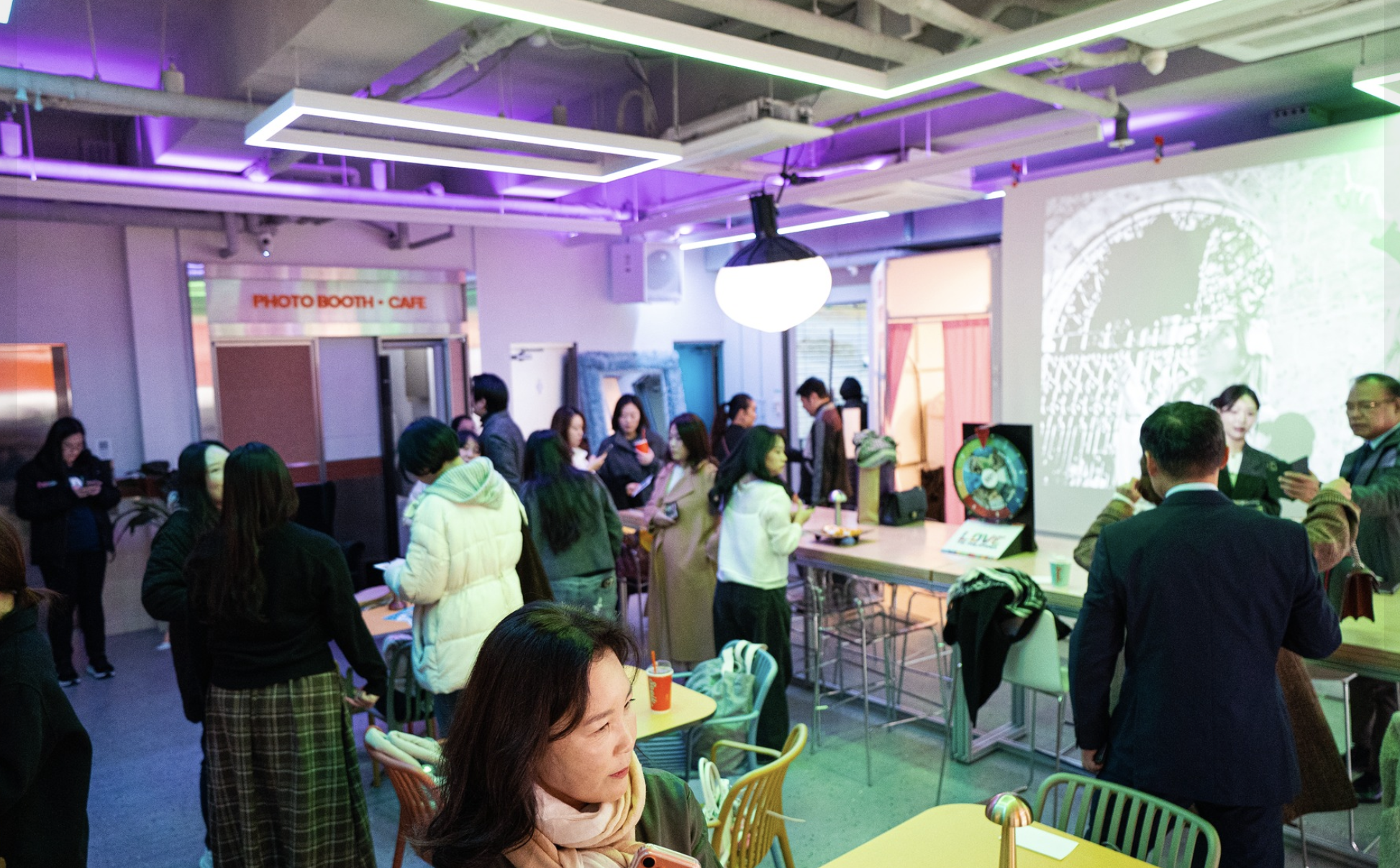 People gathered inside a modern cafe or event space with colorful lighting. Some are standing and conversing, while others are seated at tables. There is a projection on the wall and various decorations, creating a lively and social atmosphere.