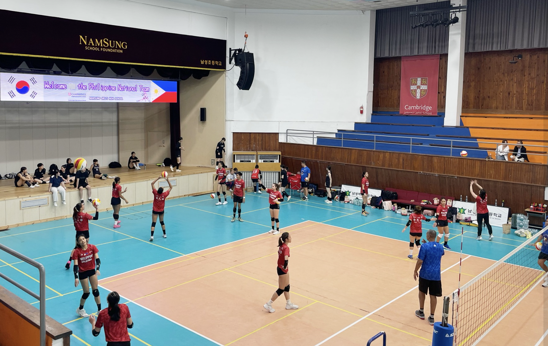 An indoor volleyball court with players warming up and practicing; spectators seated on bleachers; banners and scoreboard displayed in the background.