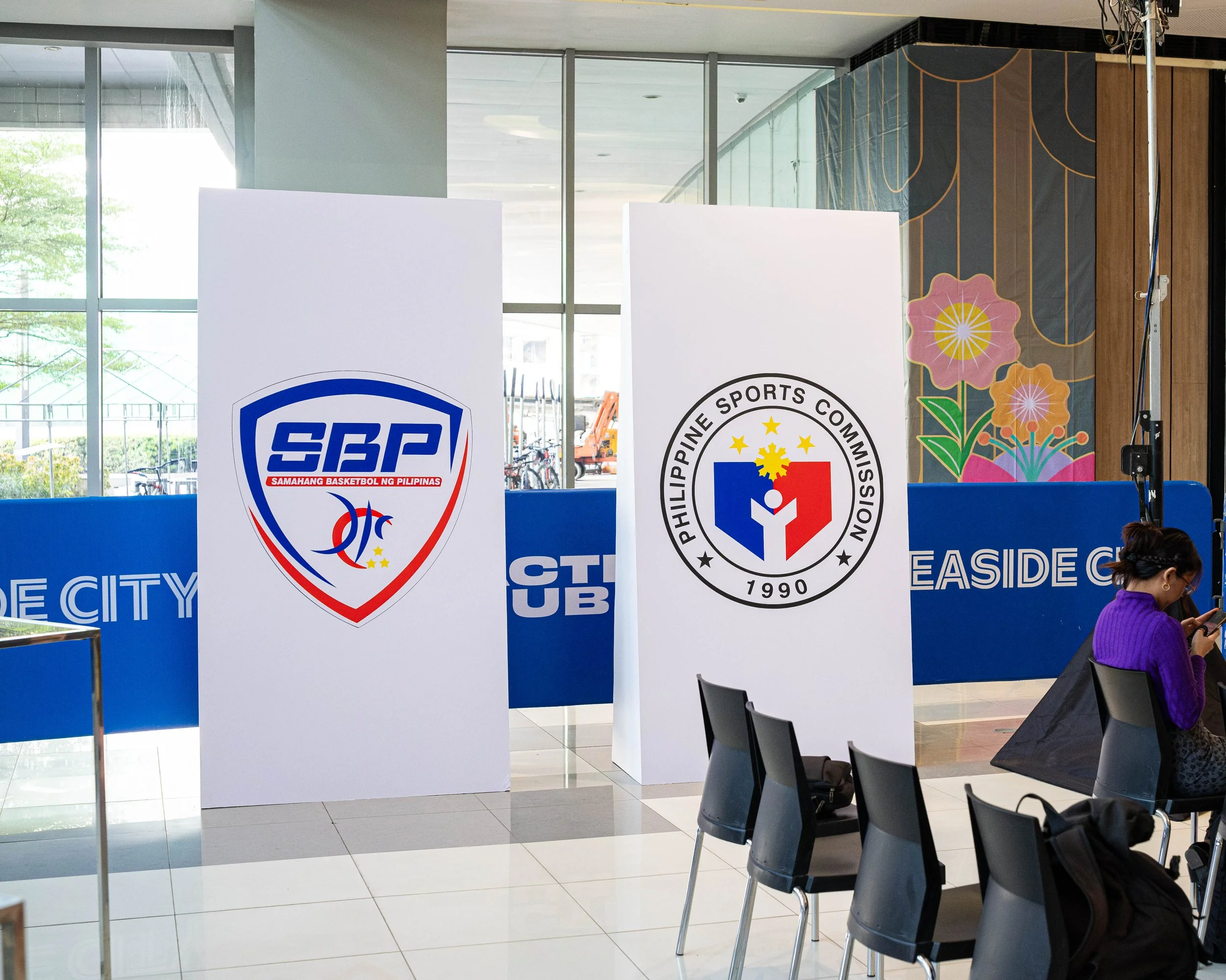 Indoor space with large windows, displaying banners for Philippine Sports Commission and SPS (Samahang Basketbol ng Pilipinas). There are chairs, a person looking at a phone, and decorative artwork with flowers on the wall.