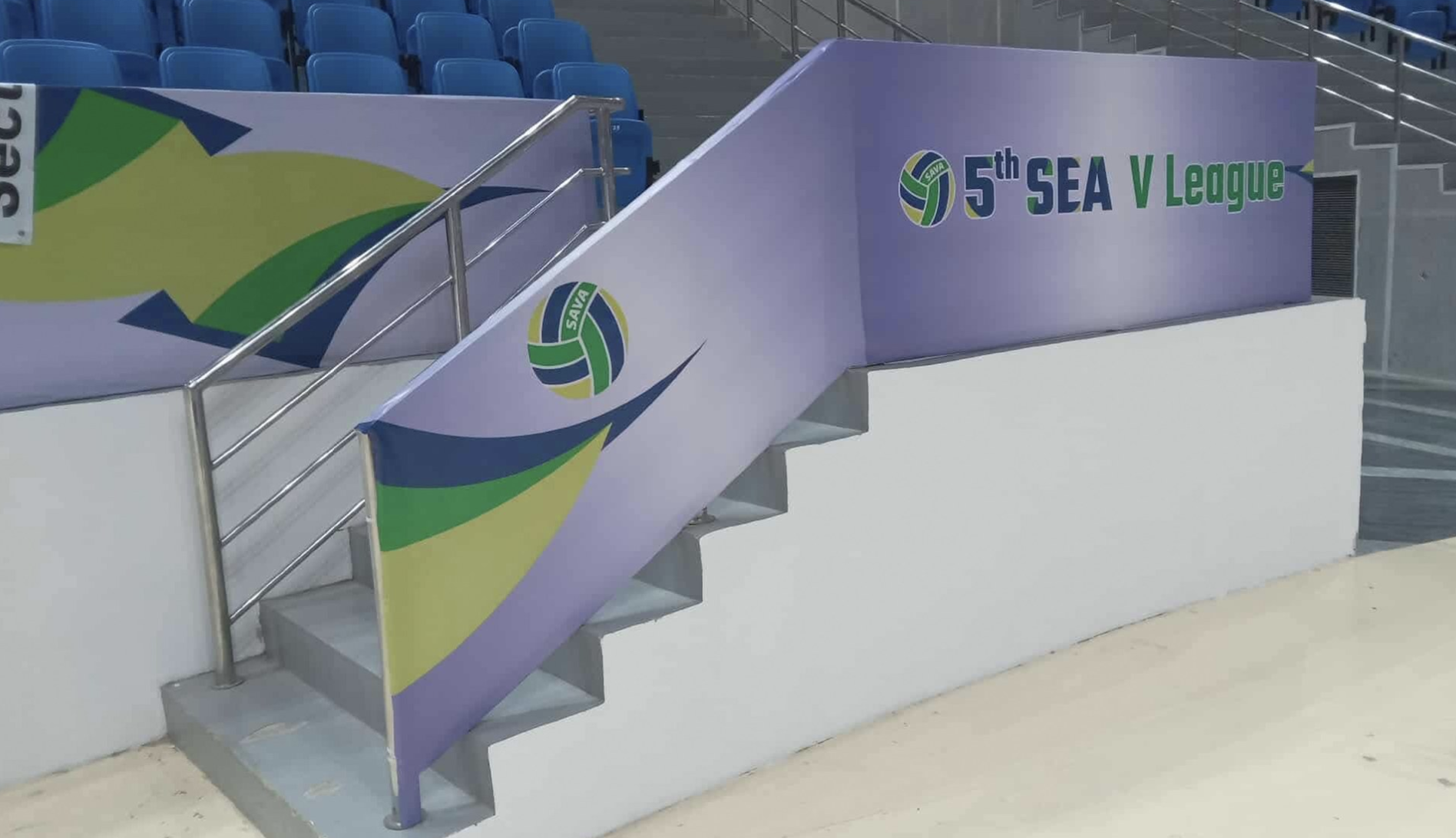 Stadium seating with blue chairs and a purple team banner for the 5th SEA V League, featuring a circular logo with green, blue, and yellow elements.