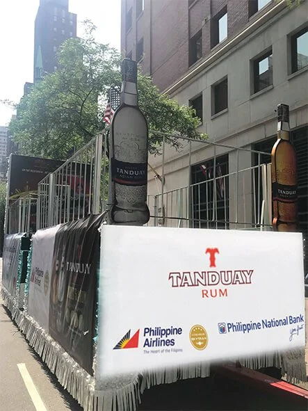 Large mural of a Tanduay Rum bottle behind a railing with Tanduay branding and logo on a white banner, along with logos of Philippine Airlines, Philippine National Bank, and other sponsors on a city street.