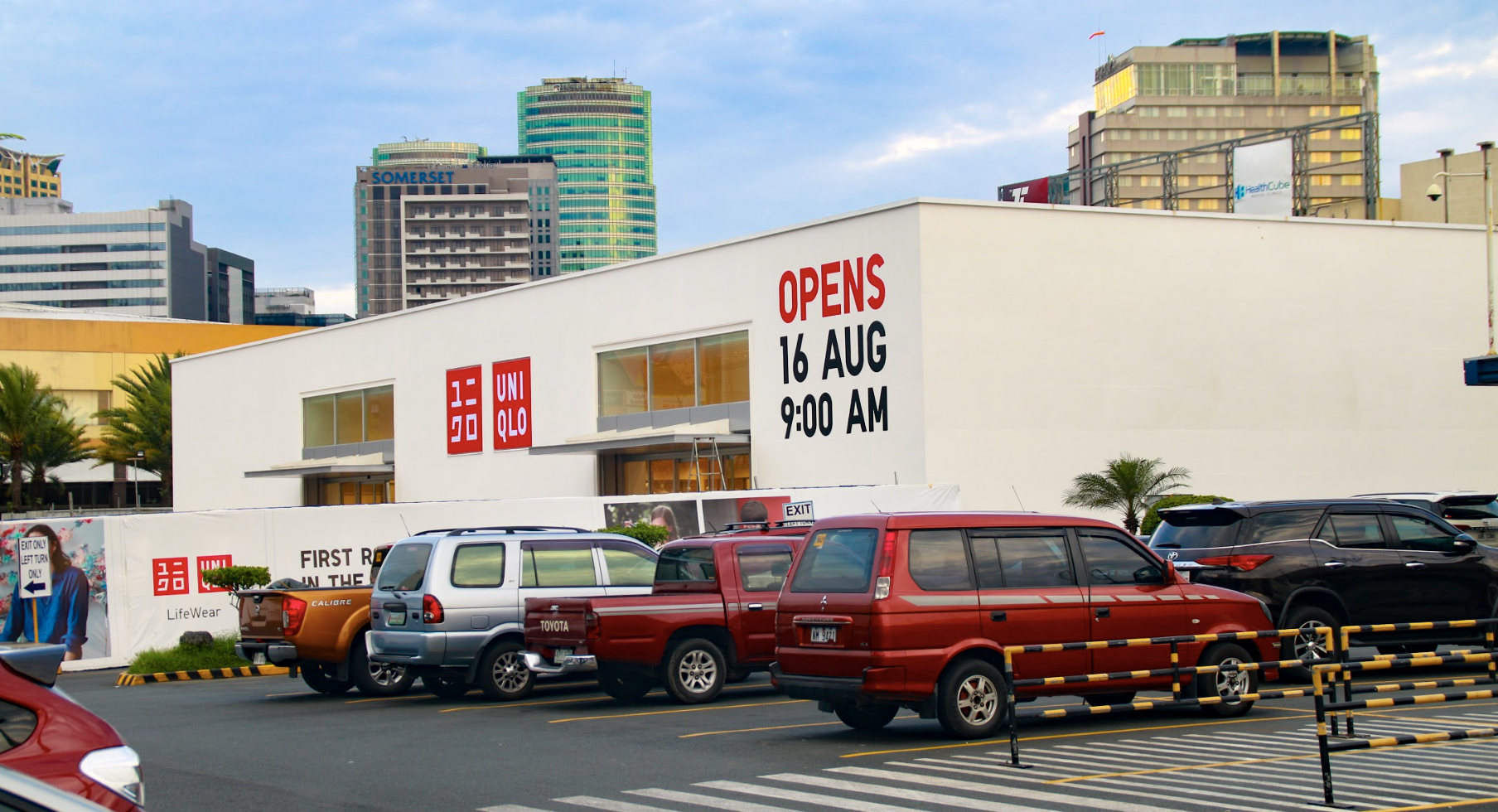 A shopping mall parking lot with several parked cars in front of a store. The store has signage indicating it will open on August 16 at 9:00 AM, featuring the UNIQLO brand logo and a sign that says 'OPENS 16 AUG 9:00 AM.' High-rise buildings can be s