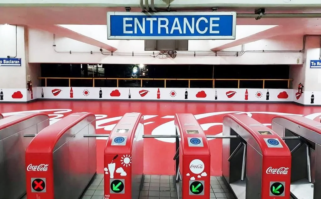 Entrance to a parking garage with red turnstiles featuring Coca-Cola branding, some with a green arrow indicating entry and one with a red cross indicating no entry. The background has a Coca-Cola themed mural with bottle icons and clouds.