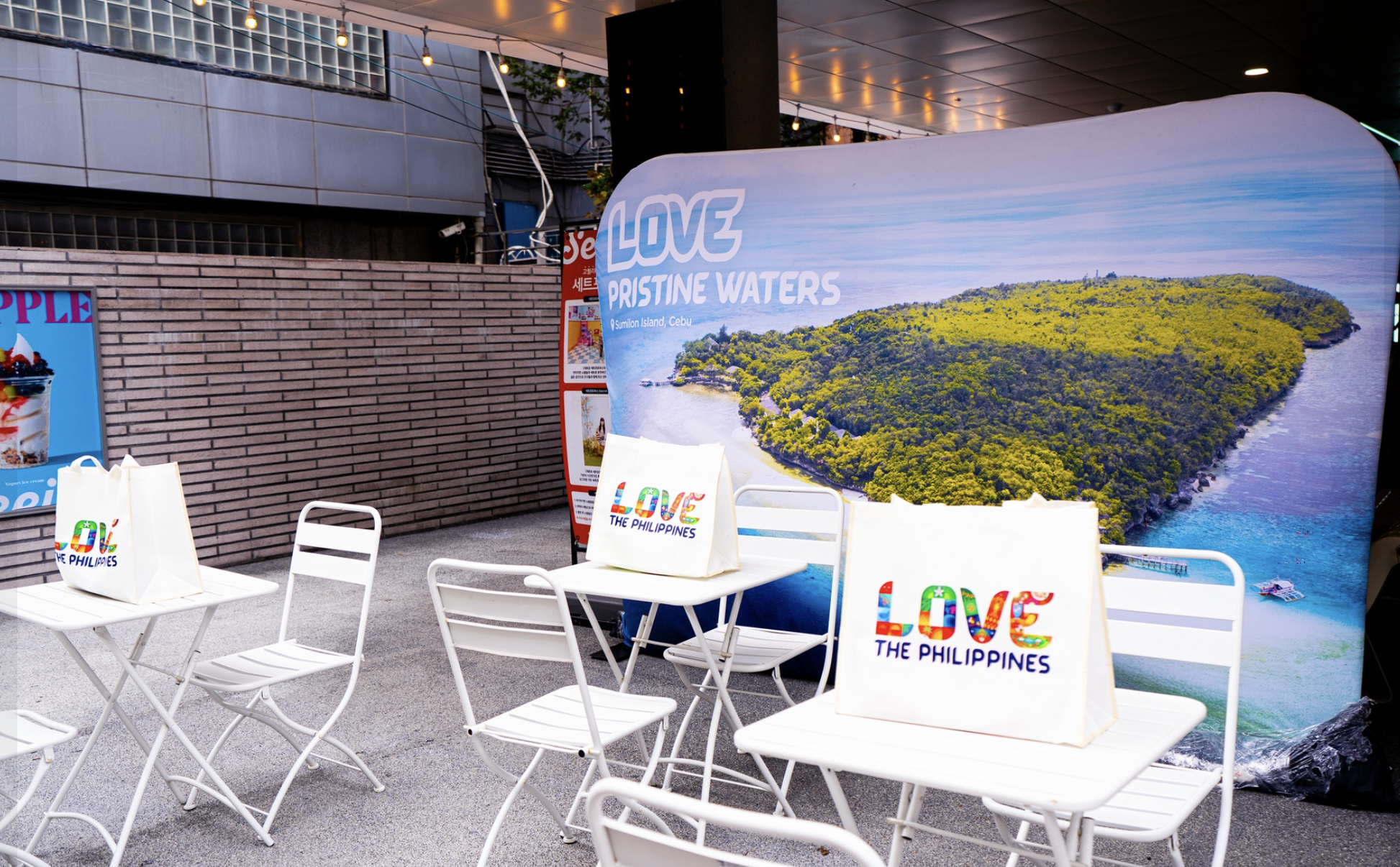 Outdoor promotional setup for love Philippines with a large banner showing a lush island surrounded by water, white chairs and tables with white bags featuring colorful Philippines flag design, and string lights overhead.