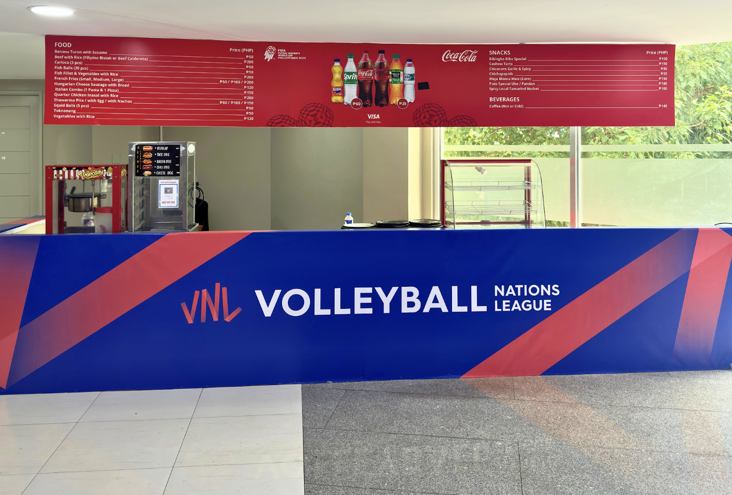 A volleyball event booth with a blue and red banner that reads "VNL Volleyball Nations League." Behind the booth, there is a red menu board with food and beverage prices and images of bottled drinks. To the left, a popcorn machine and a snack vendor 