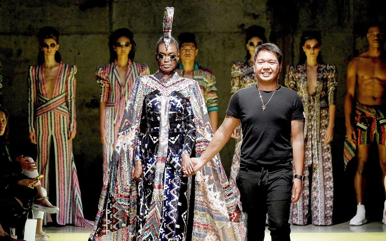 Fashion show runway scene with models wearing colorful, patterned outfits and designer walking with assistant.