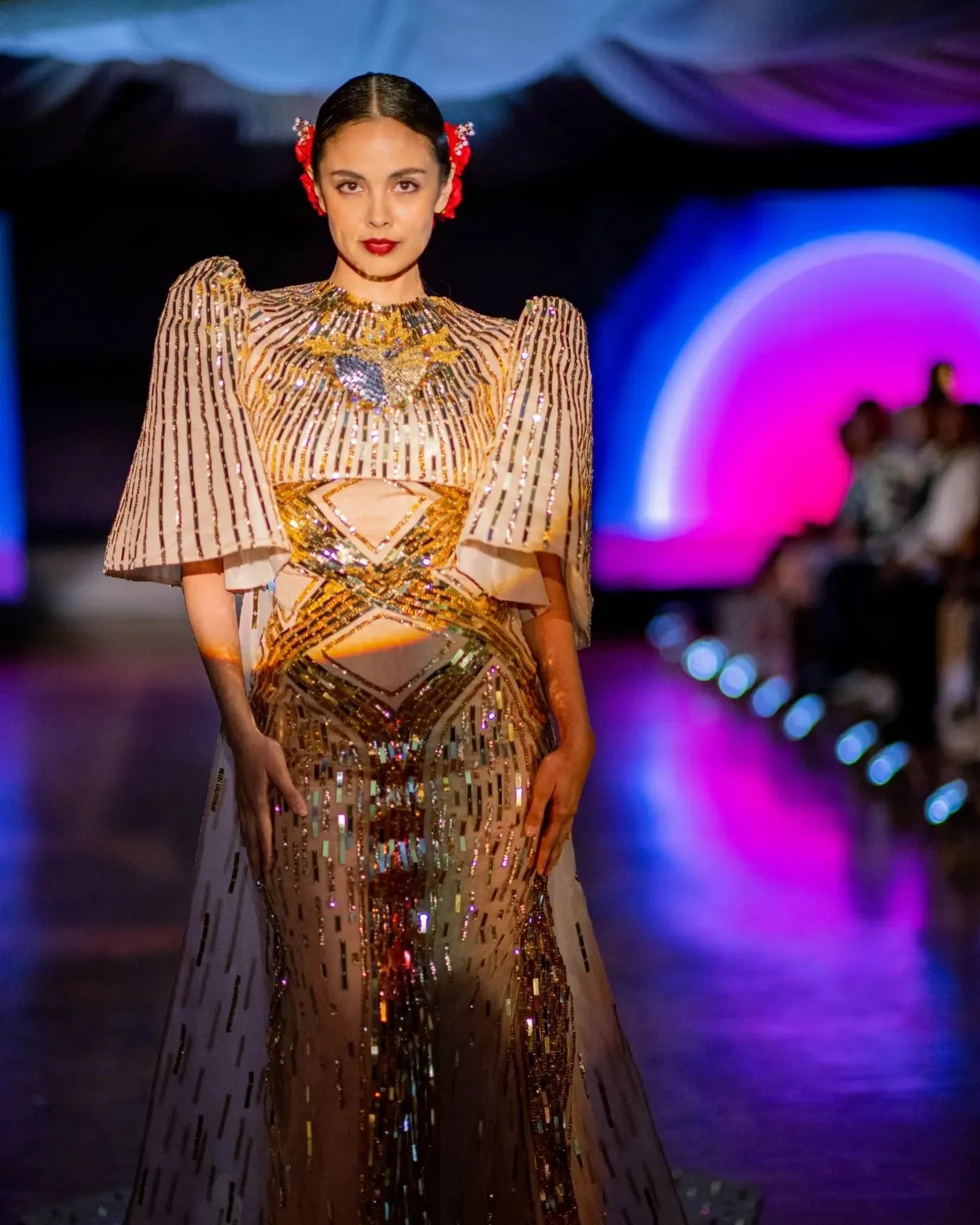 A woman walking on a runway wearing a beige and gold shimmering, intricately designed gown with puffy sleeves, adorned with sequins and beads. She has dark hair styled back with red floral accessories and wears red lipstick.