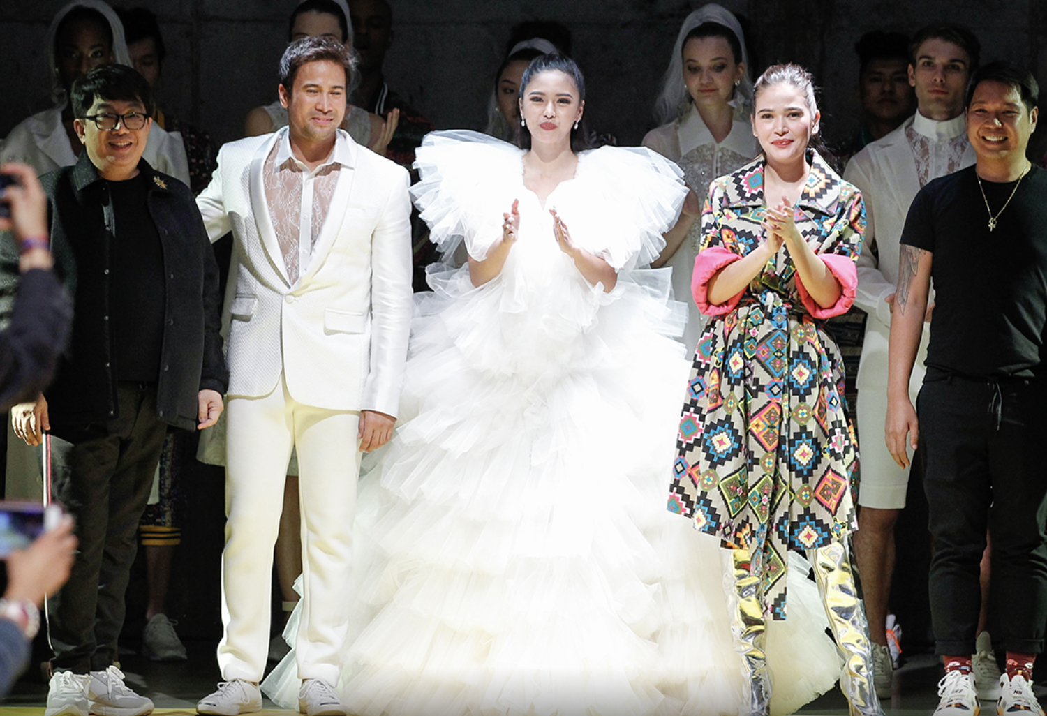 Group of people on stage at a fashion show, with a bride in a white wedding gown at center, flanked by two men and a woman in colorful clothing.
