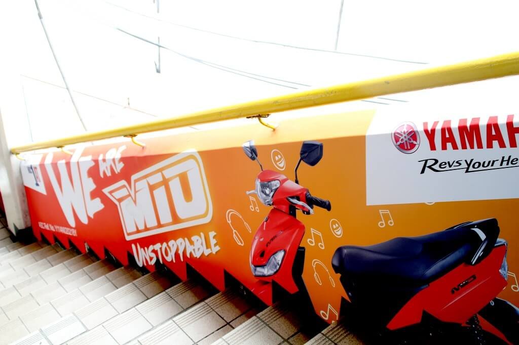 A red Yamaha scooter with black seat parked beside a colorful advertisement banner on a tiled floor. The banner features a scooter illustration, Yamaha logo, and the words "We Are Mito Unstoppable" with musical note icons.