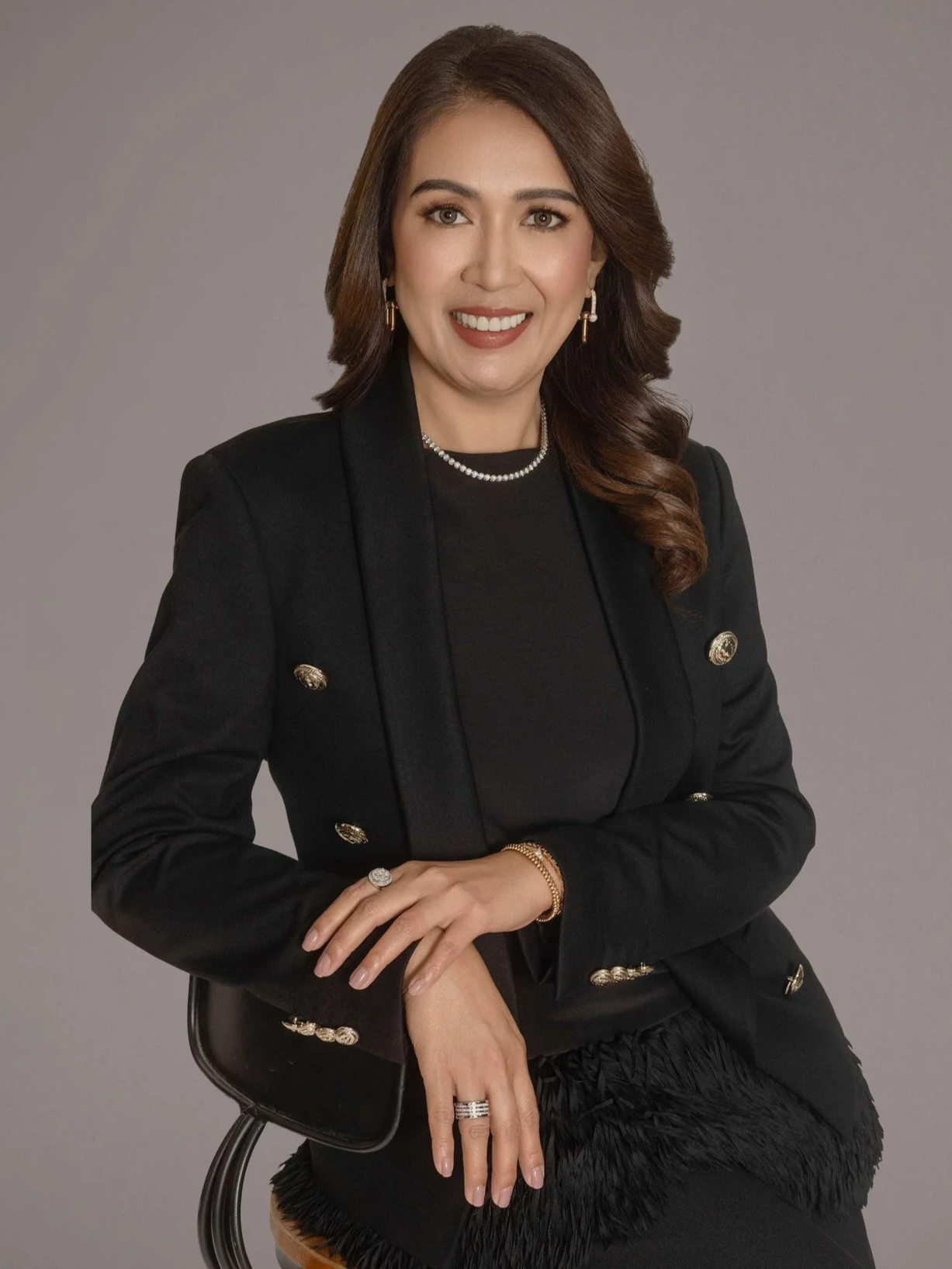 Professional woman with shoulder-length brown hair, wearing a black blazer with gold buttons, a black top, and jewelry, sitting on a black chair against a neutral background.