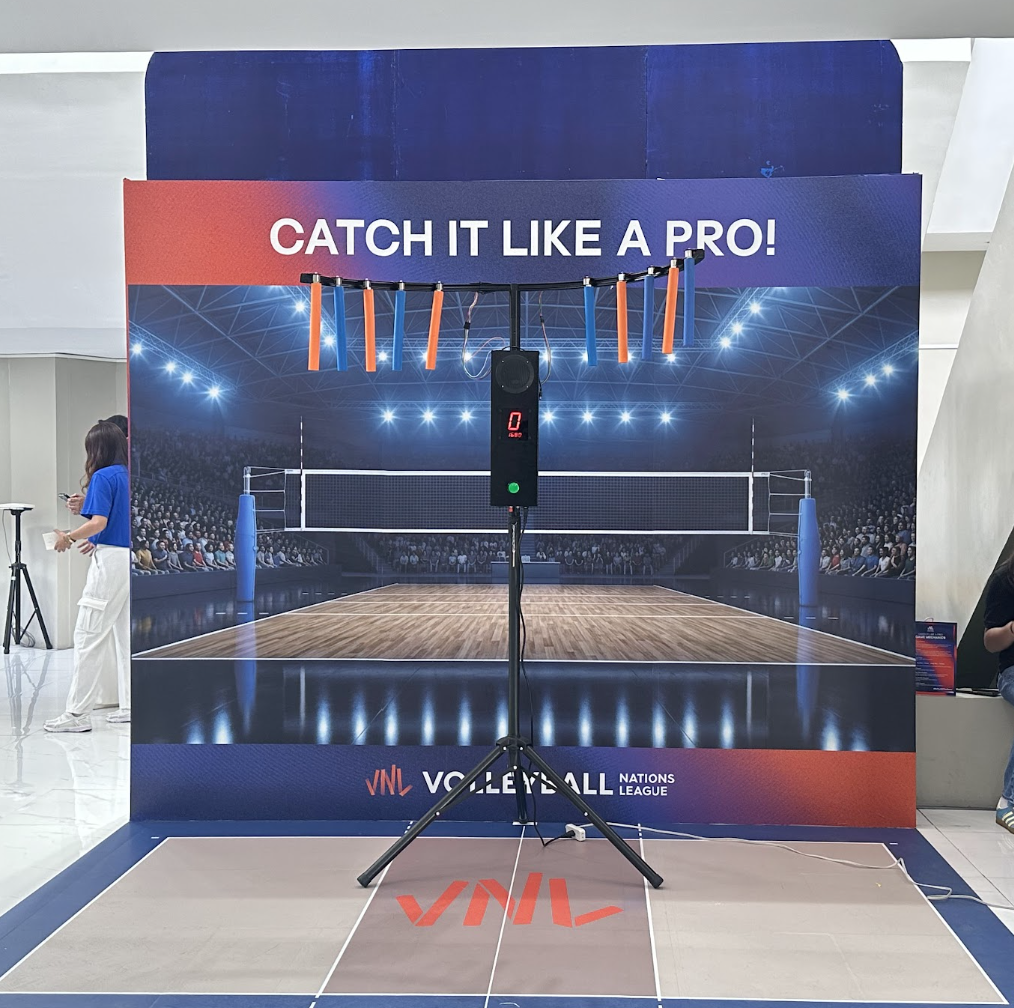 A volleyball court with a sign that reads "Catch It Like a Pro!" and a digital timer on a stand in front of the court. The background features a digital image of an indoor arena filled with a crowd.