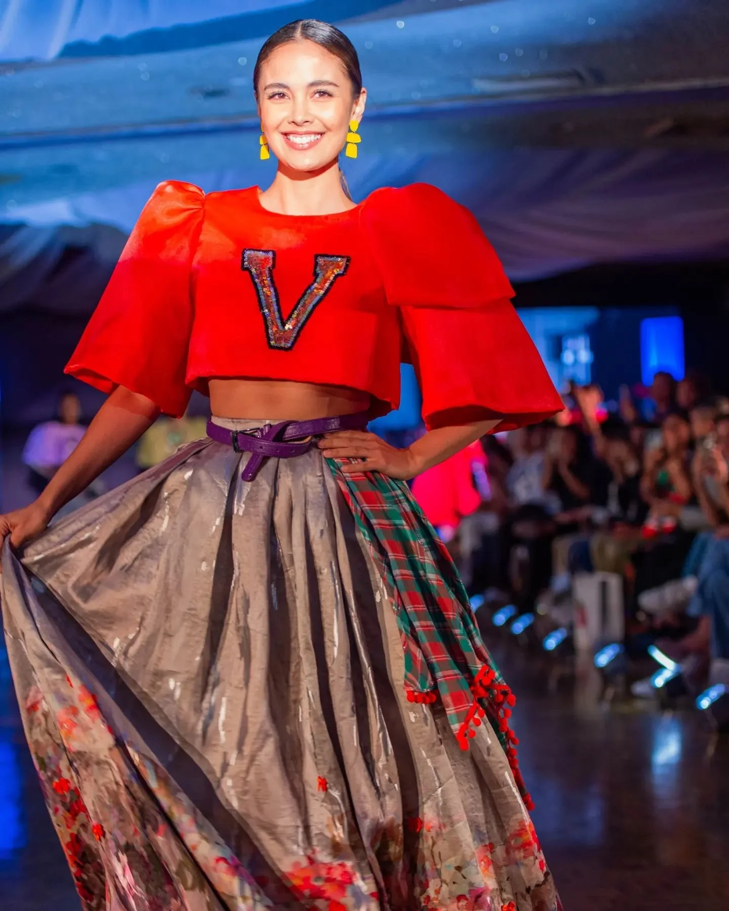 A woman walking down a runway wearing a colorful, patterned full-length skirt with a metallic finish, a red crop top with puffed sleeves, and yellow earrings, smiling with her hand on her hip, in a fashion show setting.
