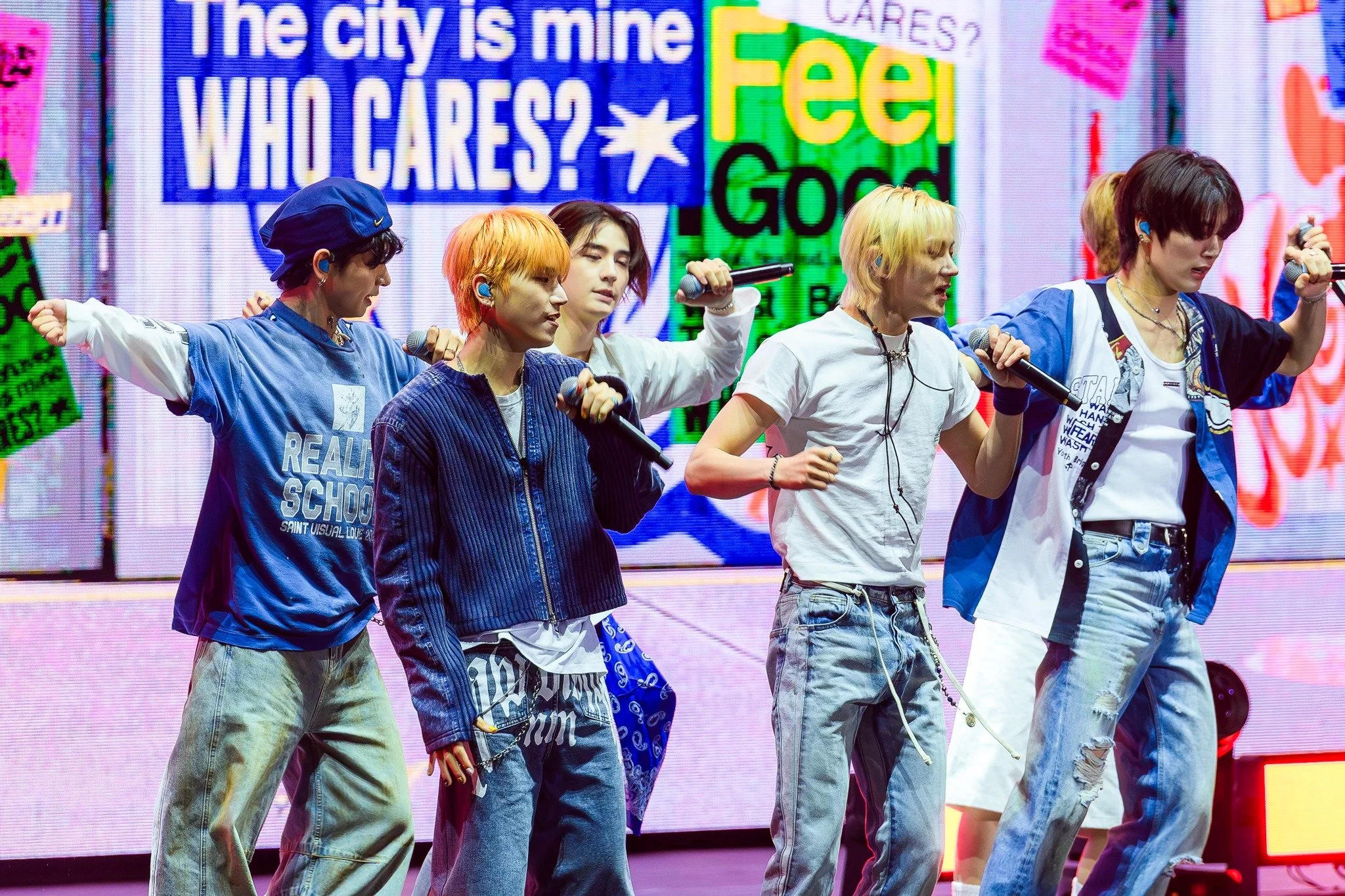 A group of six young men standing on stage, dressed in casual streetwear, holding microphones, with colorful signage and a digital screen behind them.