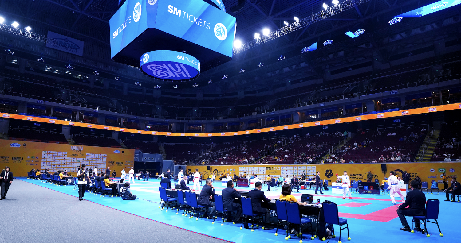 Indoor arena hosting a karate competition with competitors, officials, and spectators, large electronic scoreboard displays SM Tickets, and yellow banners with event information.